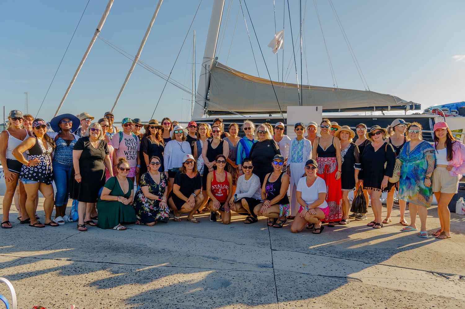A large group of women posing on a dock in front of a sailing catamaran at golden hour