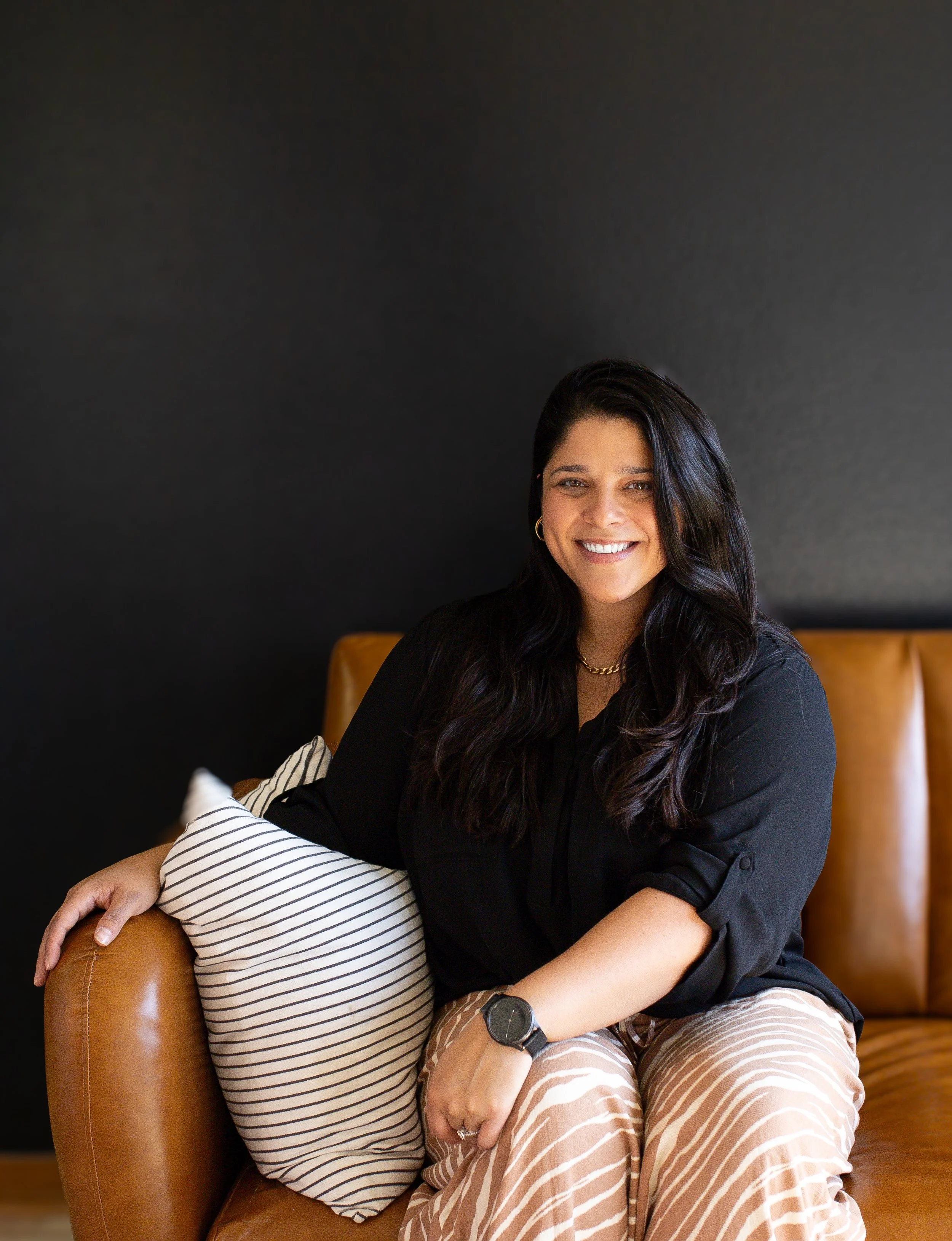 a picture of MMFT Student Therapist Vanessa Gonsalves sitting on a brown leather couch smiling at the camera