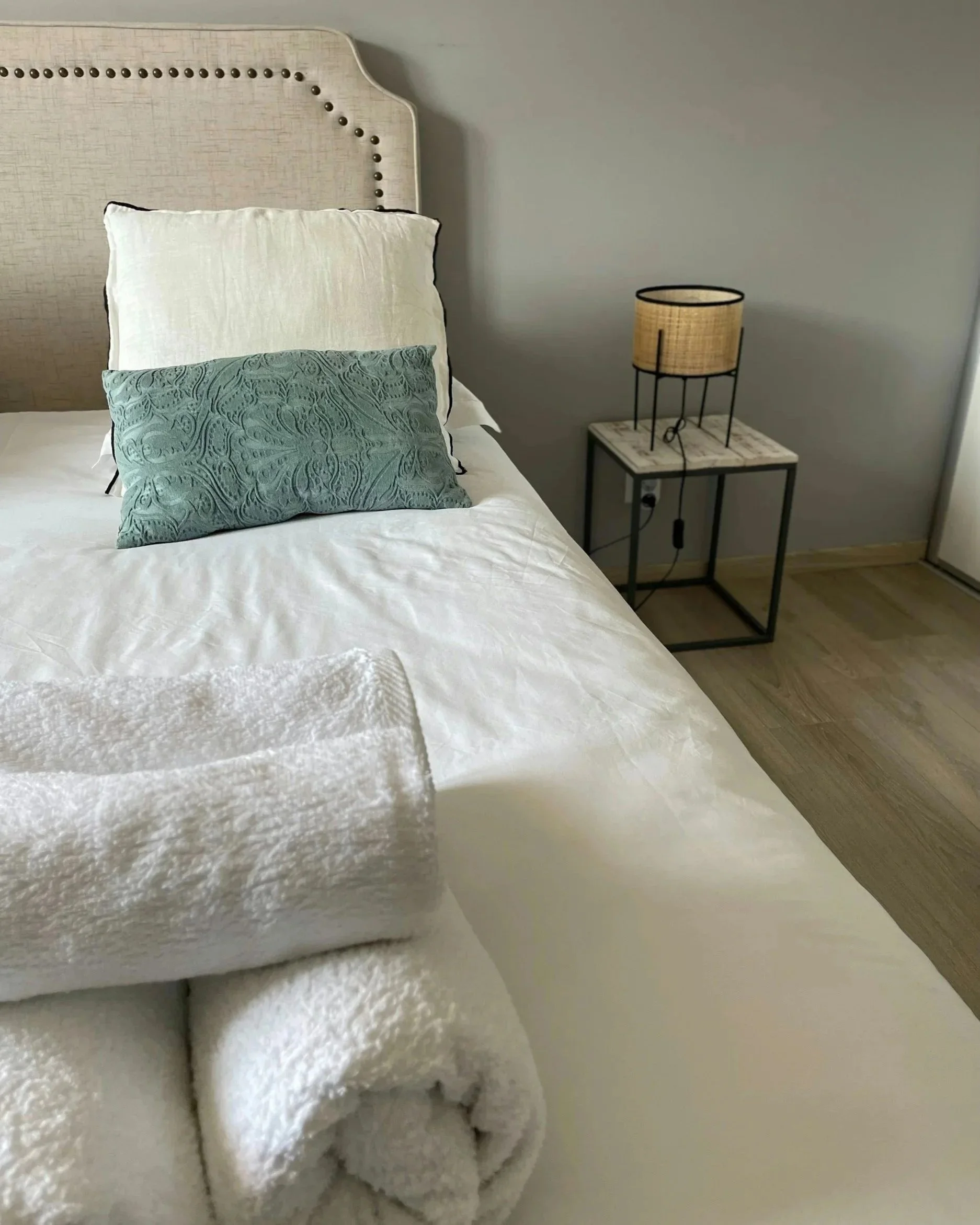 Neatly made bed with white linens, white and patterned teal pillows, and rolled towels beside a bedside table and lamp.