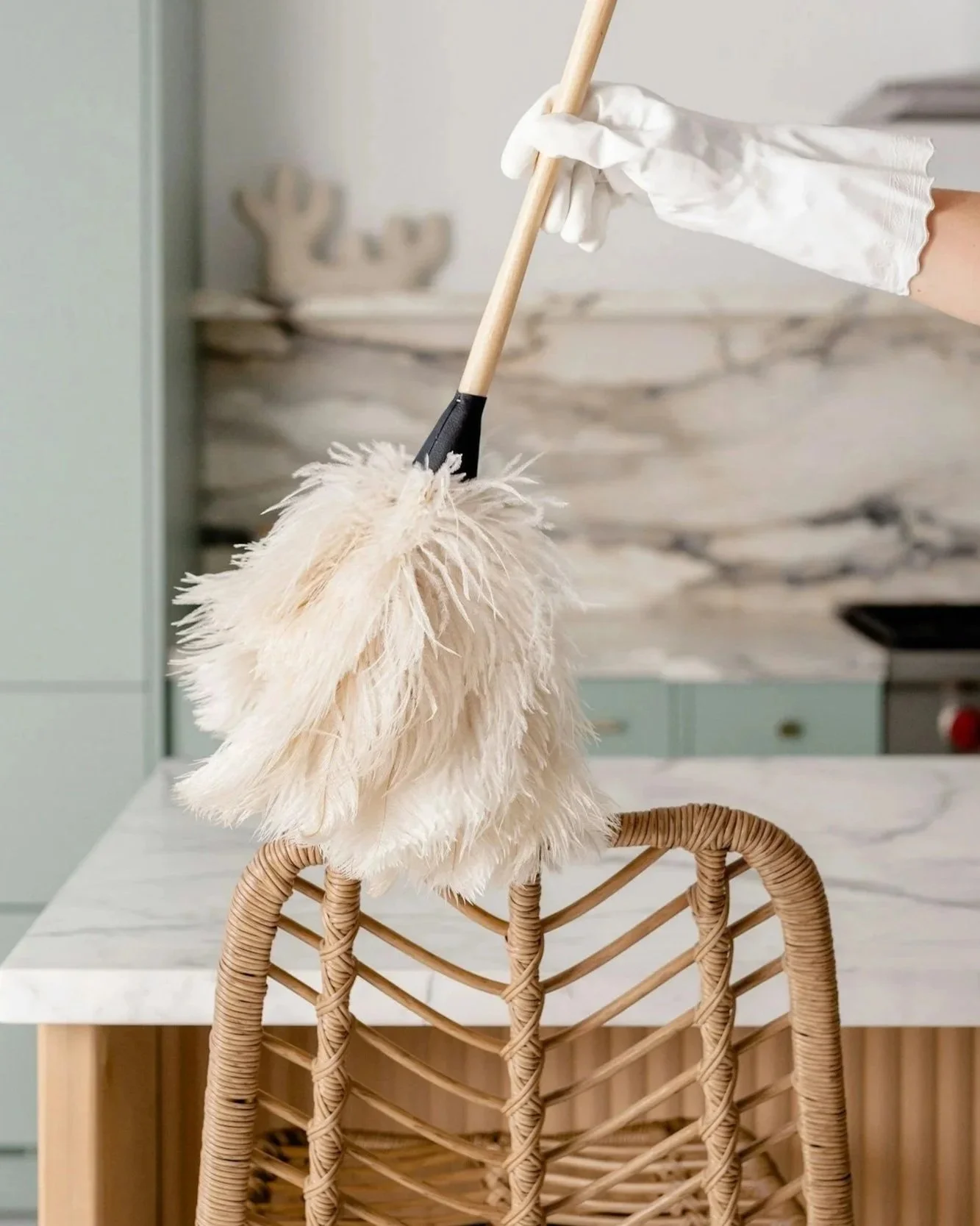 Gloved hand dusting a wicker chair with a fluffy feather duster in a bright kitchen