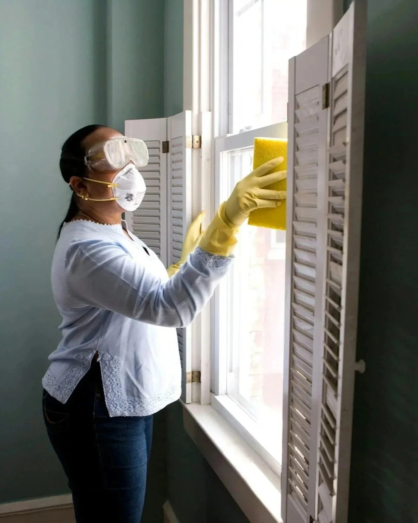 Woman wearing protective goggles, face mask, and gloves cleans a window with a yellow sponge.