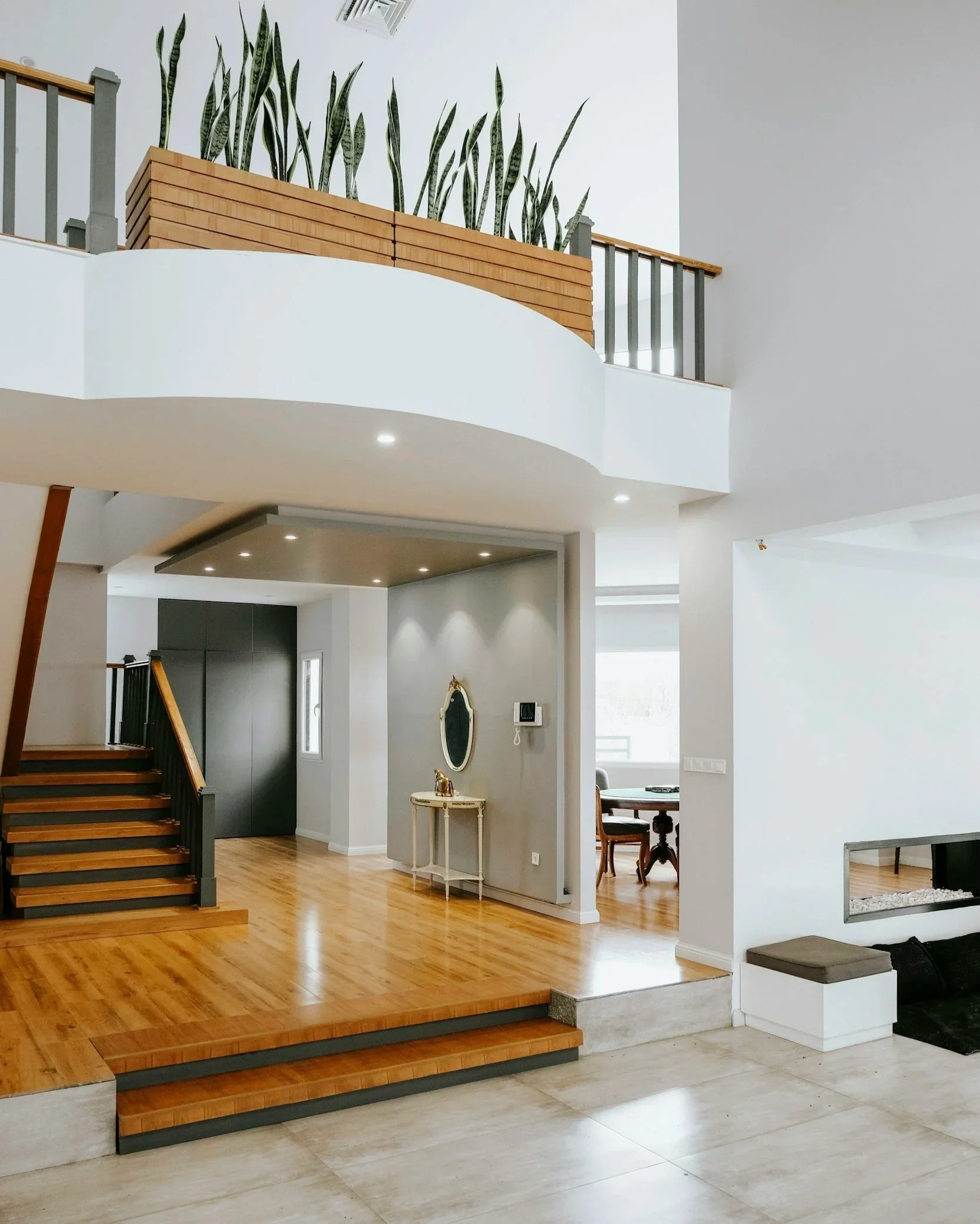 Modern open-concept home interior with wood staircase, mezzanine railing, indoor snake plants, polished hardwood floors, and fireplace