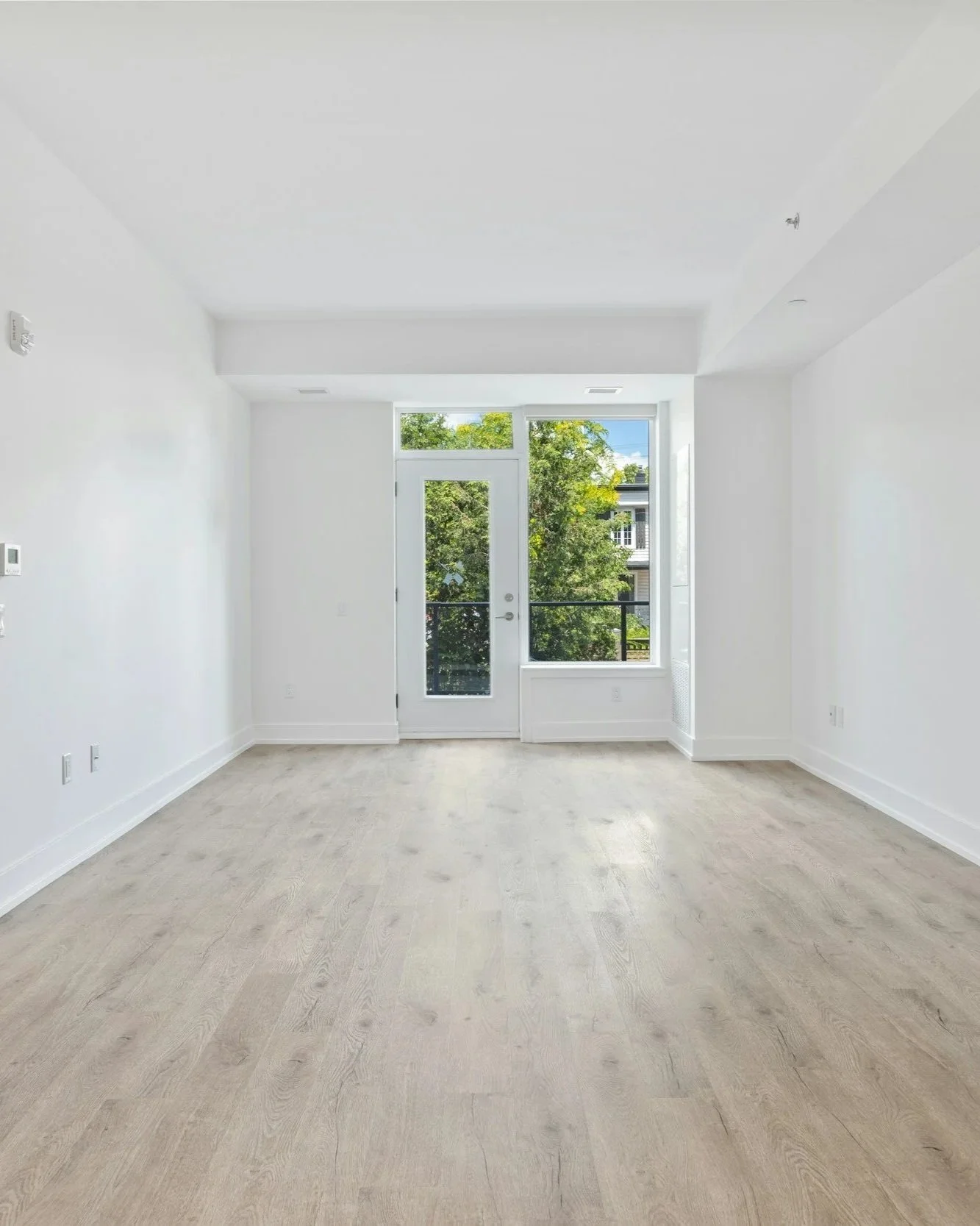 Bright empty white room with light wood flooring, glass door, large window, and recessed ceiling lights