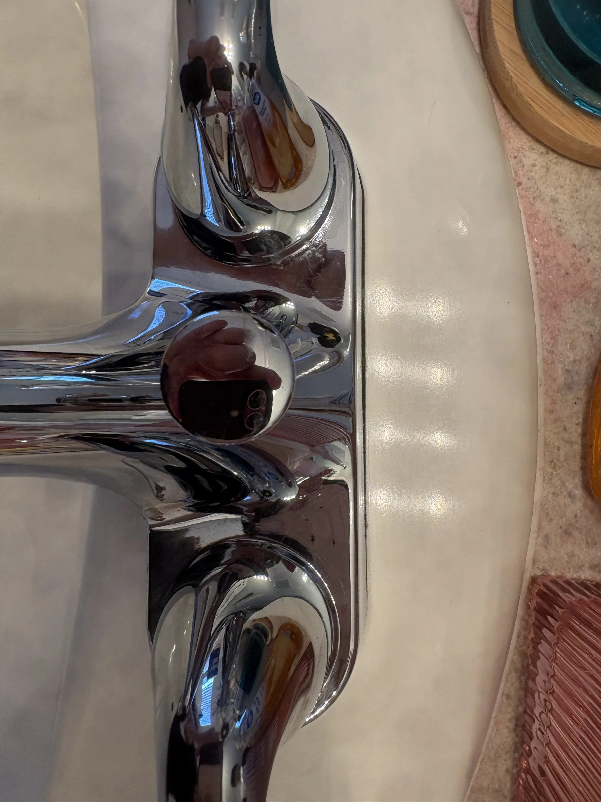 Close-up of a shiny chrome faucet reflecting a person taking a photo, beside a white sink and wooden countertop.