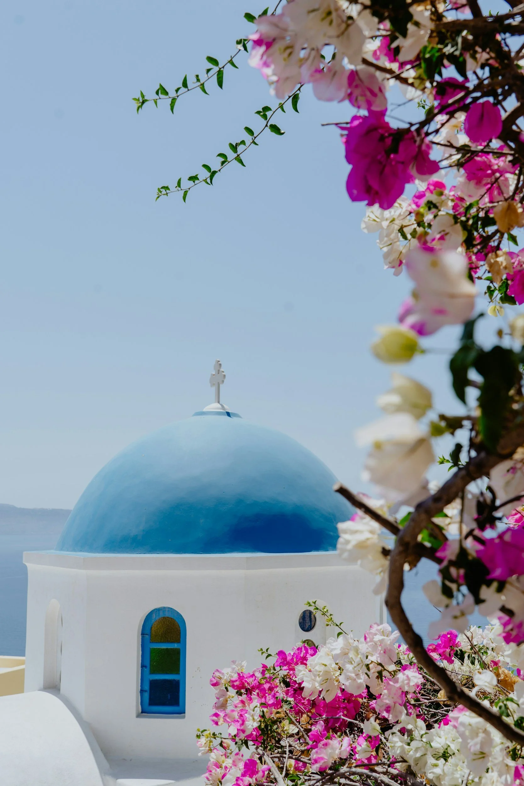 Whitewashed Greek Orthodox church with a blue dome and cross, framed by pink and white bougainvillea under a clear sky
