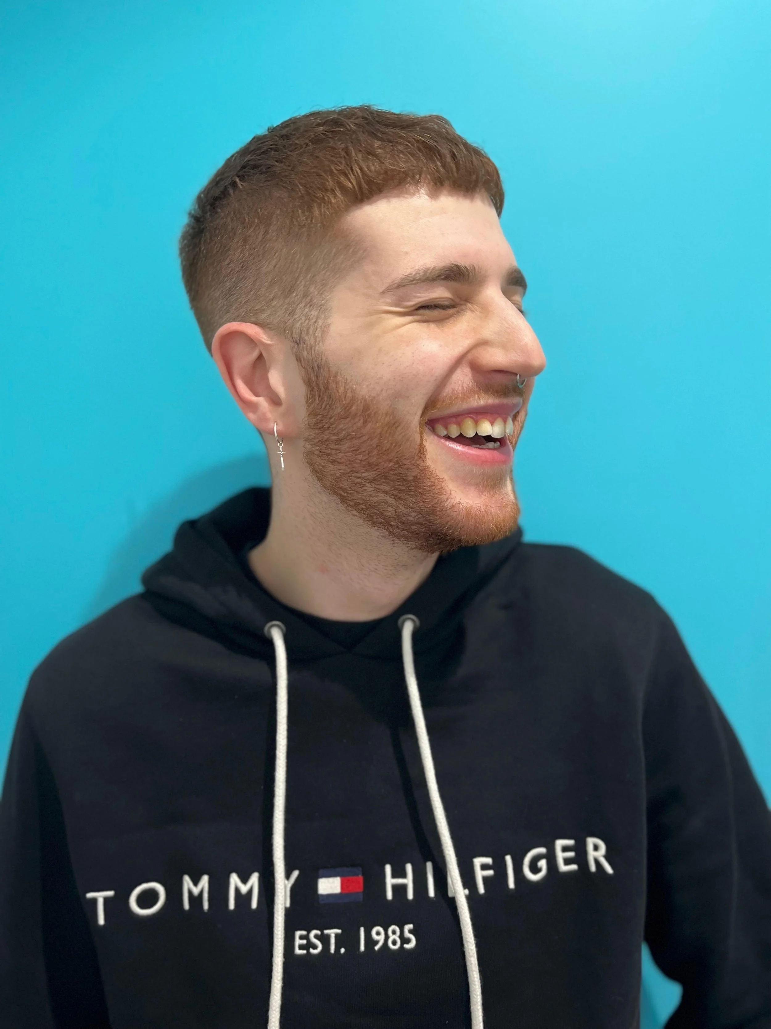 Smiling bearded man with earrings wearing a black Tommy Hilfiger hoodie against a bright blue background