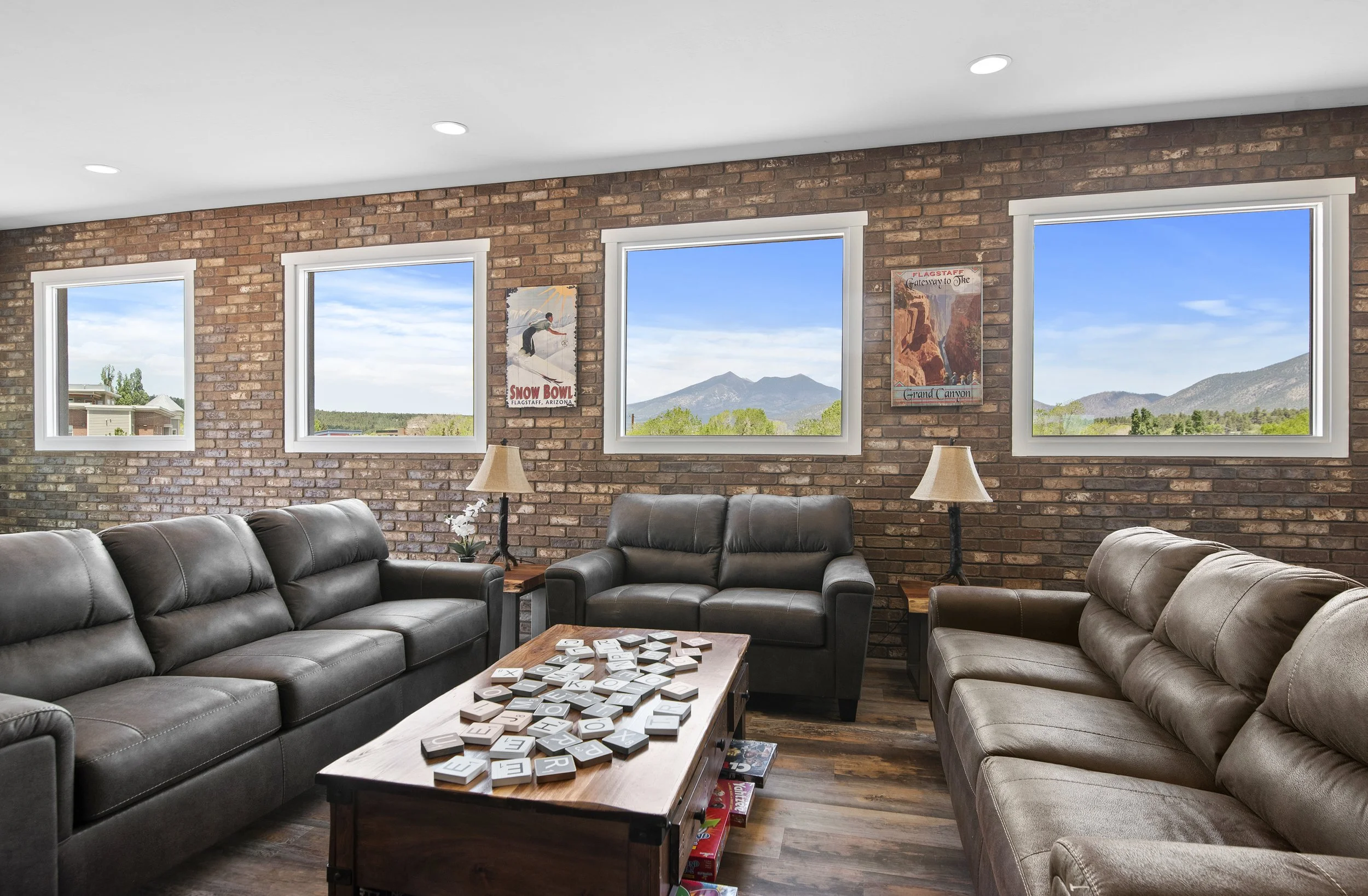 Living room with three black leather couches, a wooden coffee table with scattered letter tiles, a brick wall with three windows showing mountains and blue sky, and vintage travel posters on the wall.