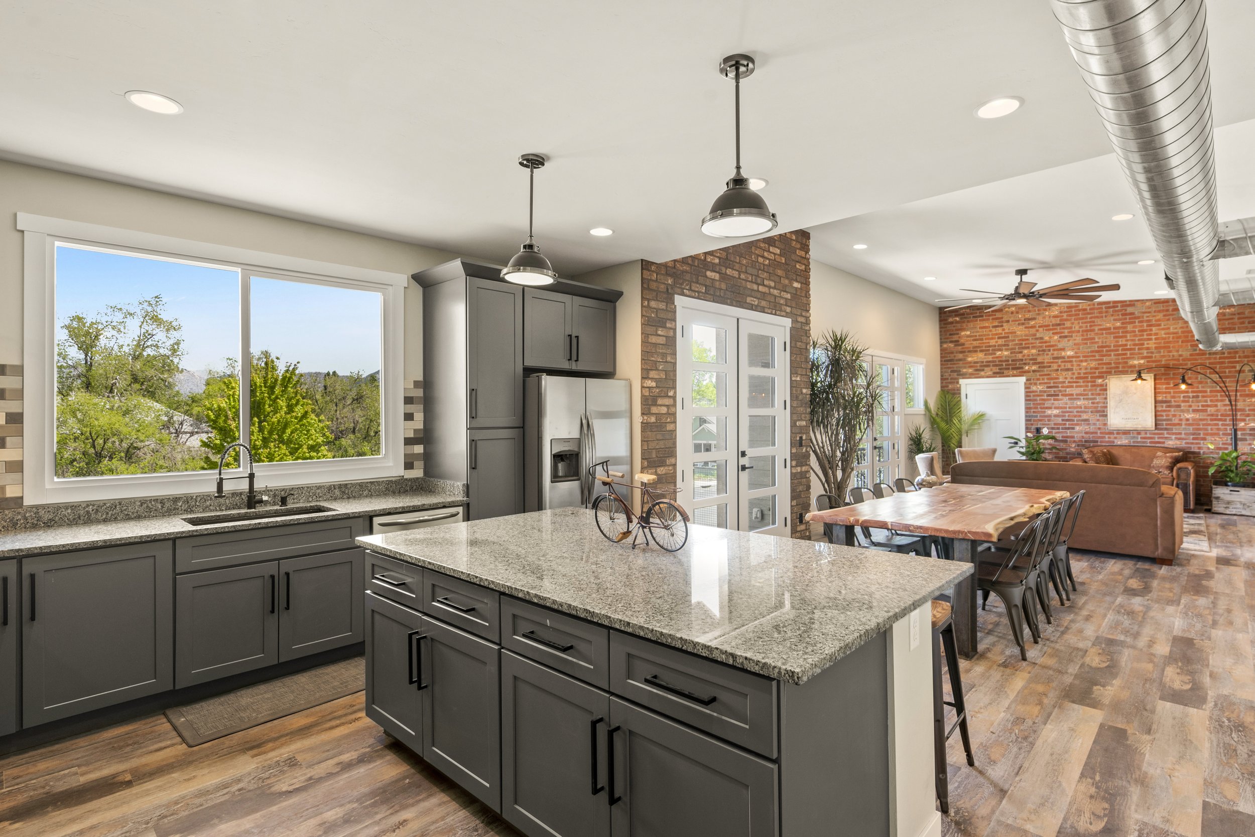 A modern kitchen with gray cabinets, a granite countertop island, a stainless steel refrigerator, and large windows looking out onto green trees. The kitchen opens into a living area with a brick accent wall, a ceiling fan, and a dining table.