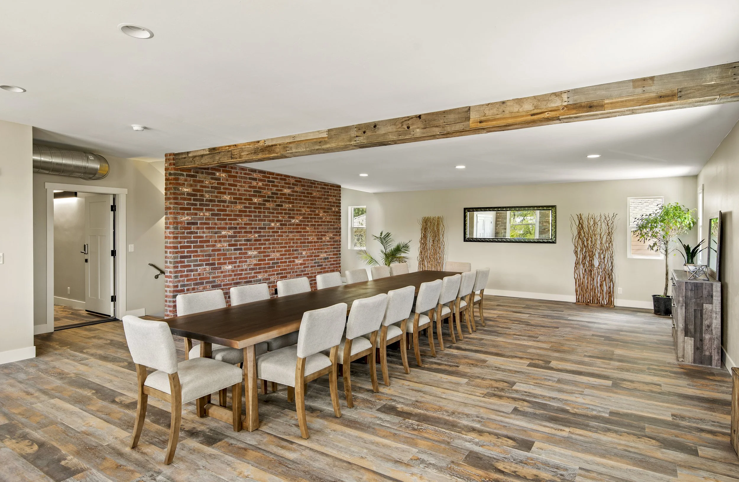 A modern dining room featuring a long wooden table surrounded by cream upholstered chairs, a brick accent wall, minimalist decor including tall dried plants and greenery, and a large mirror on the far wall. The room has wood flooring and a white ceiling with recessed lighting.