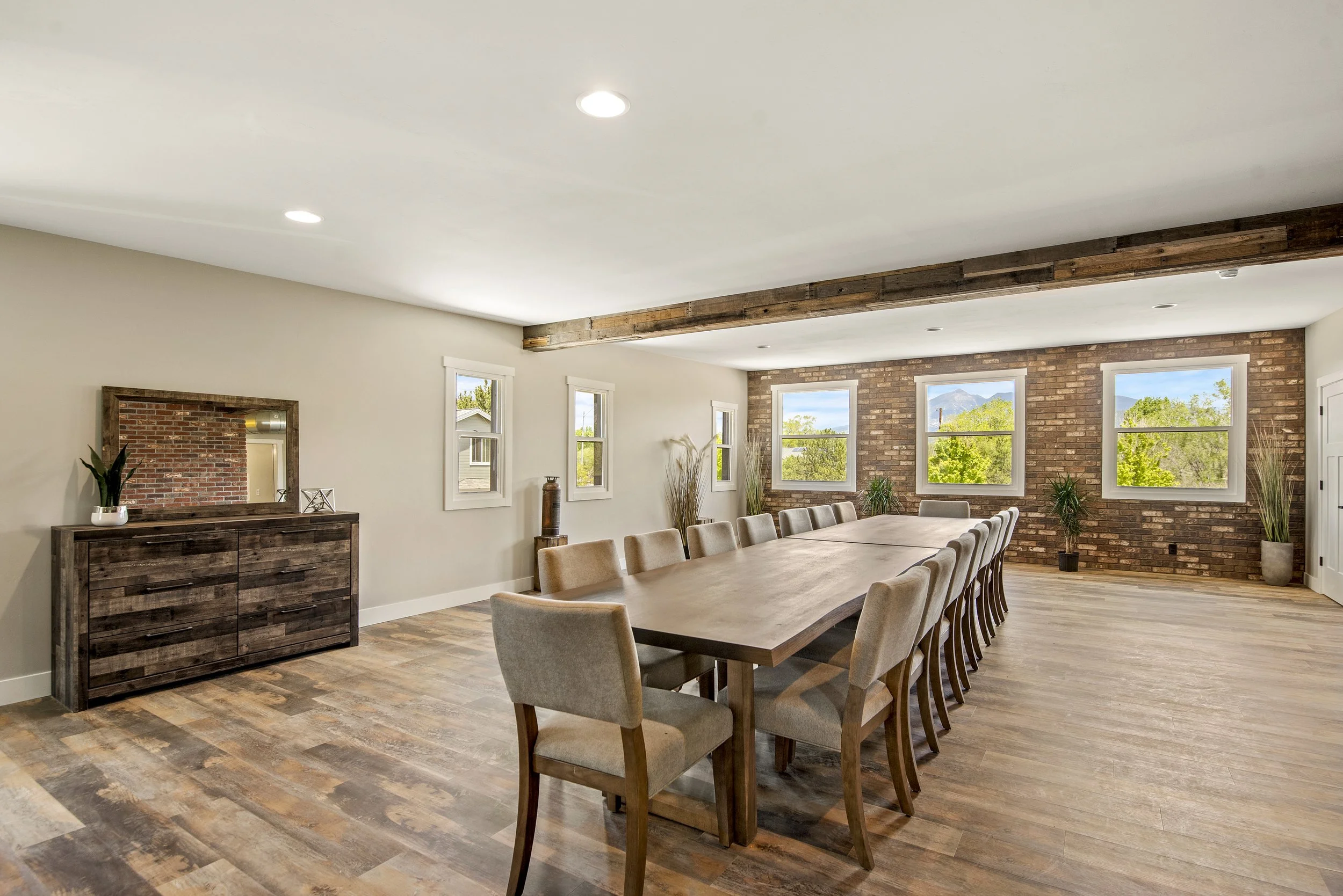 A modern dining room with a long wooden table surrounded by beige upholstered chairs, a brick accent wall with several large windows, and a ceiling beam made of reclaimed wood, with hardwood flooring and minimal decorative plants.