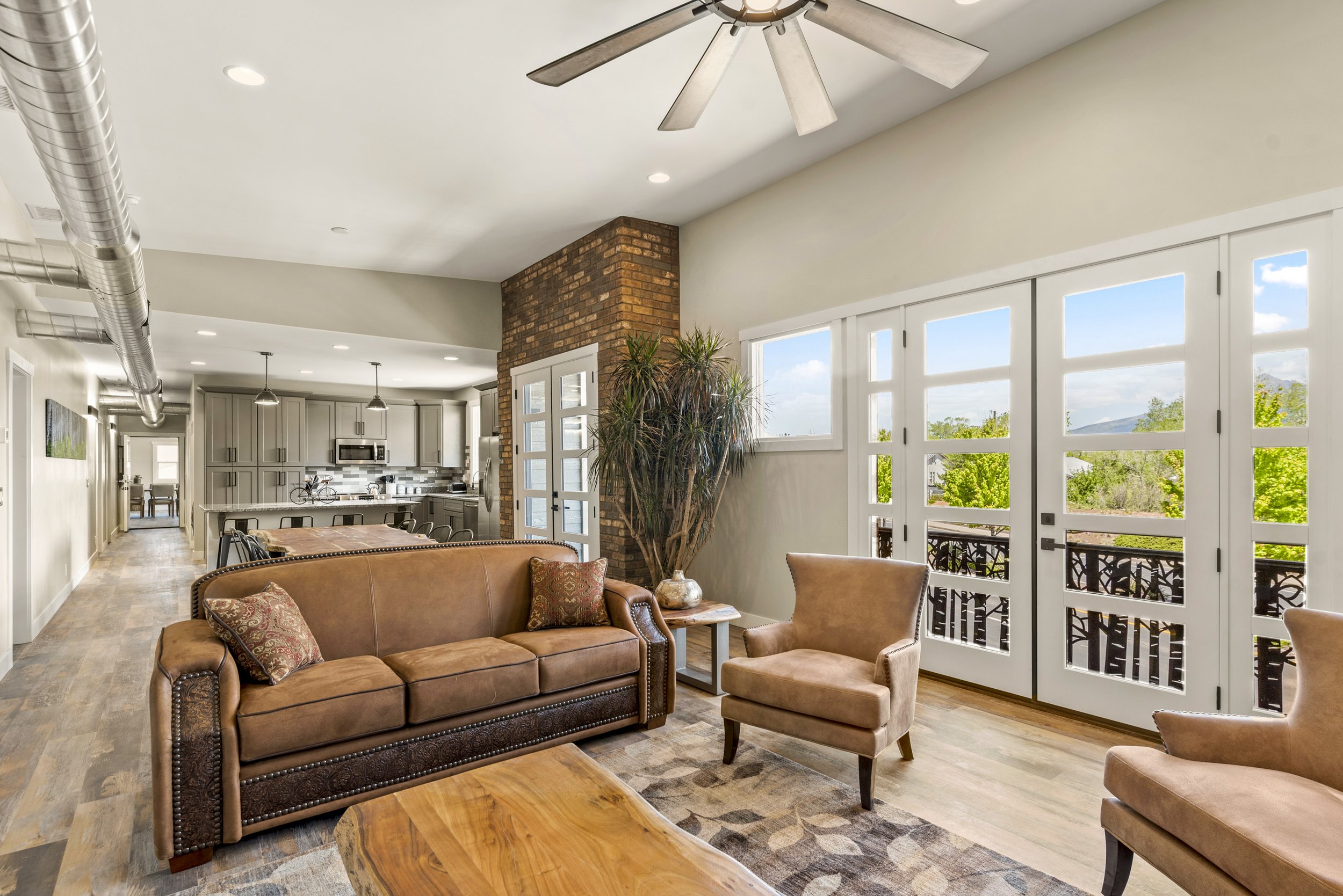 Bright living room with large windows, brick column, ceiling fan, beige walls, and leather sofa with pillows, open to kitchen area with cabinets, appliances, and dining table.