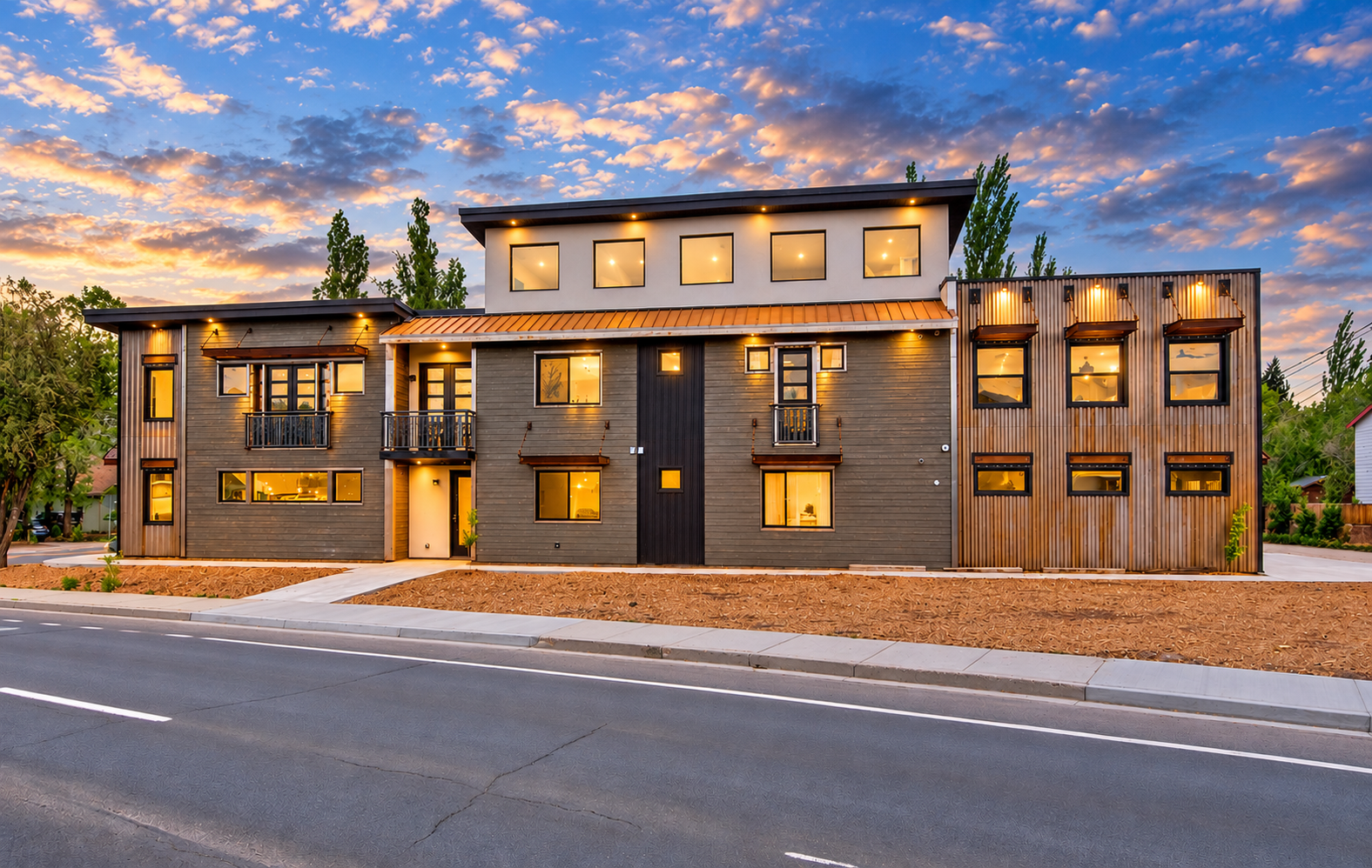 Modern multi-story residential building with wooden exterior, illuminated windows, and a sidewalk in front, during sunset.