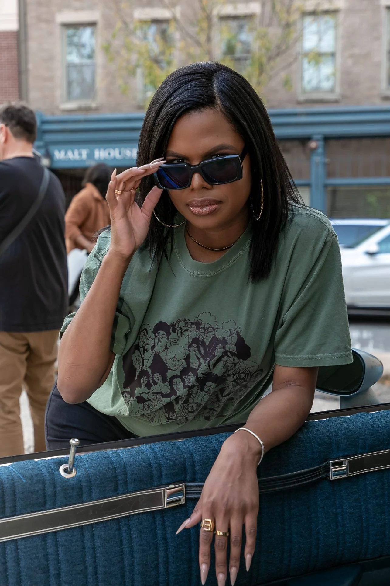 Woman wearing sunglasses and a graphic T-shirt seated in a blue vintage car on a city street