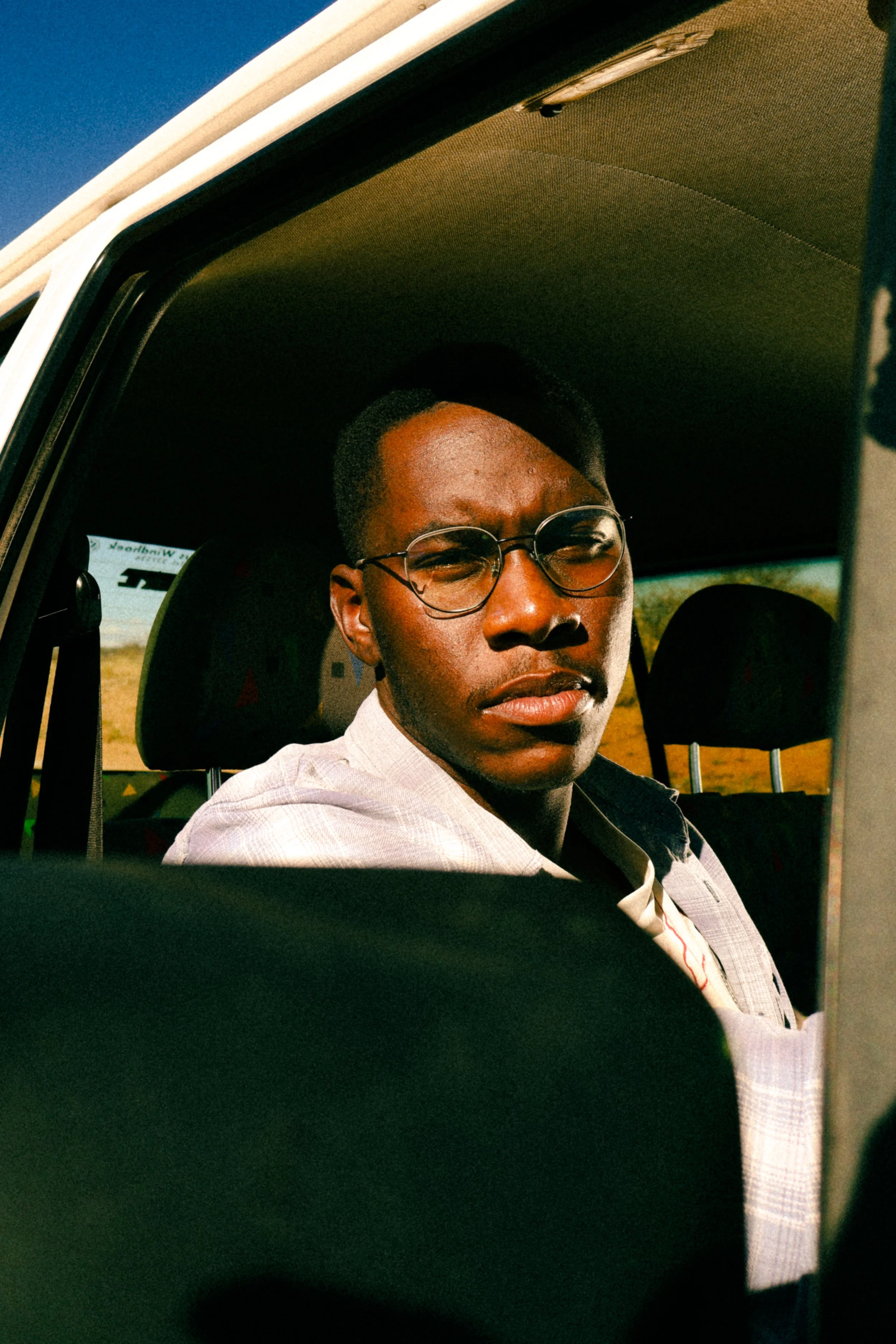 An editorial portrait photograph of Eleazar Mwakipesile looking out of the window of his car at the camera taken by Kim Kambinda in Windhoek, Namibia.