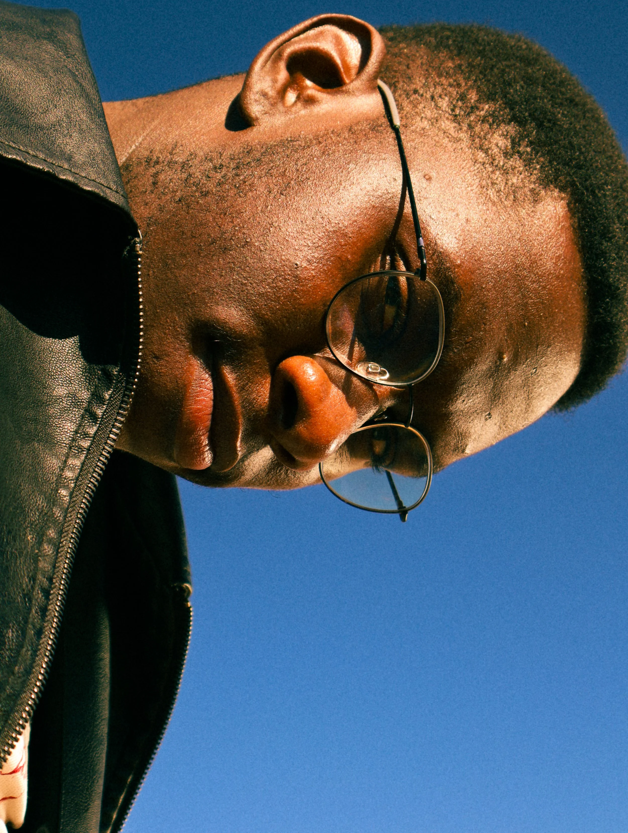 An editorial portrait photograph of Eleazar Mwakipesile looking down at the camera with the sky behind him taken by Kim Kambinda in Windhoek, Namibia.