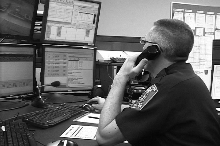 An ambulance dispatcher sitting at a desk with multiple computer monitors, speaking on a phone, with paperwork and office equipment around.