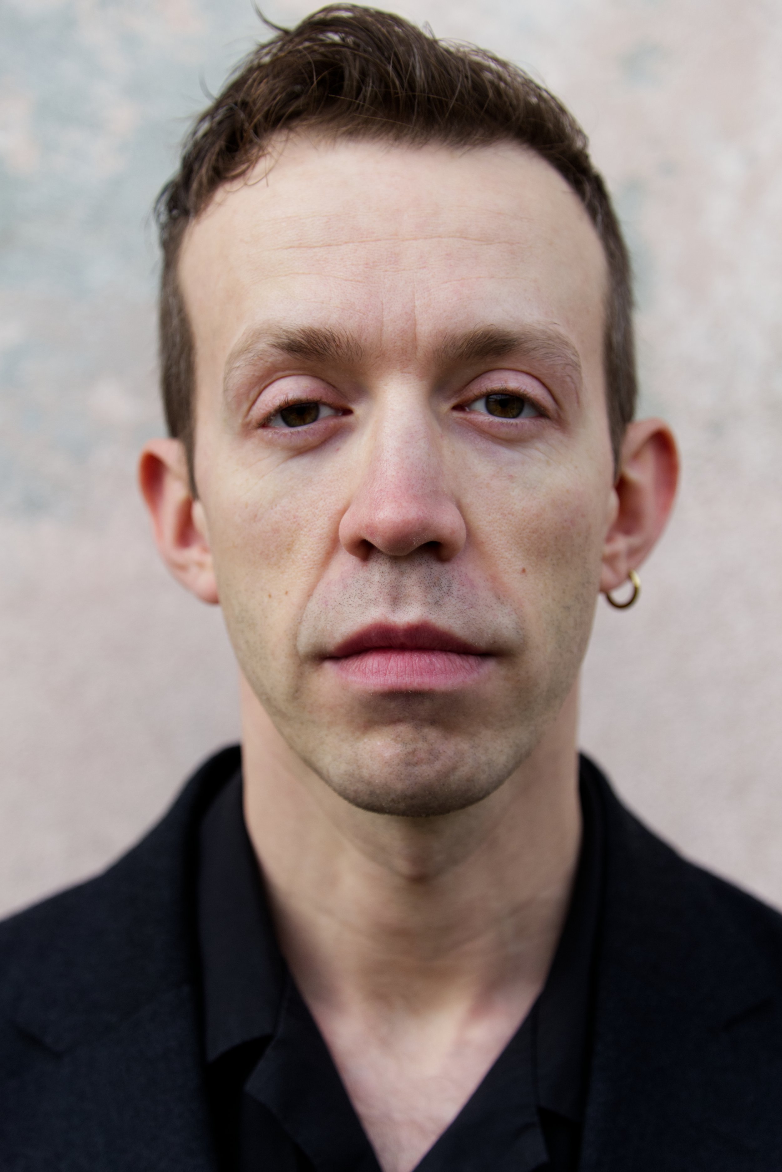 Close-up of a young man's face with short, dark hair, light skin, wearing a black shirt, and an earring in his left ear, against a light, neutral background. It is an actor mugshot of Rudi Grieser.