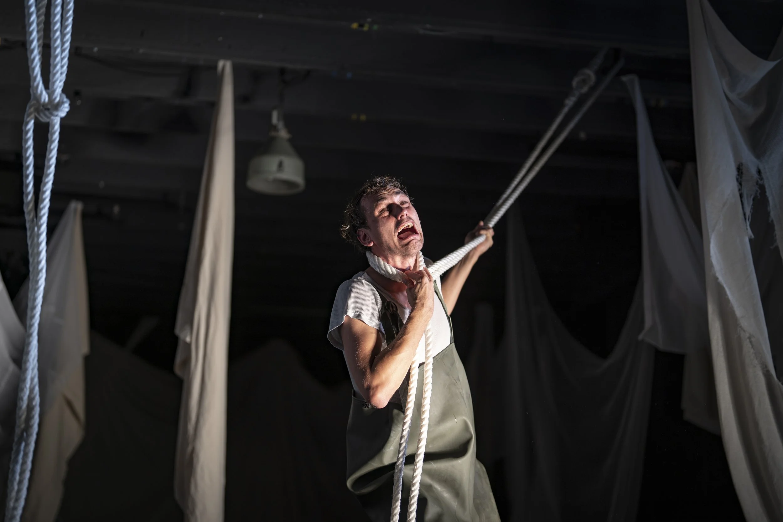 Person pulling on a thick rope in a dark space, surrounded by ropes and hanging fabric, with a  pained expression. It is an image of Hugo Tiedje.