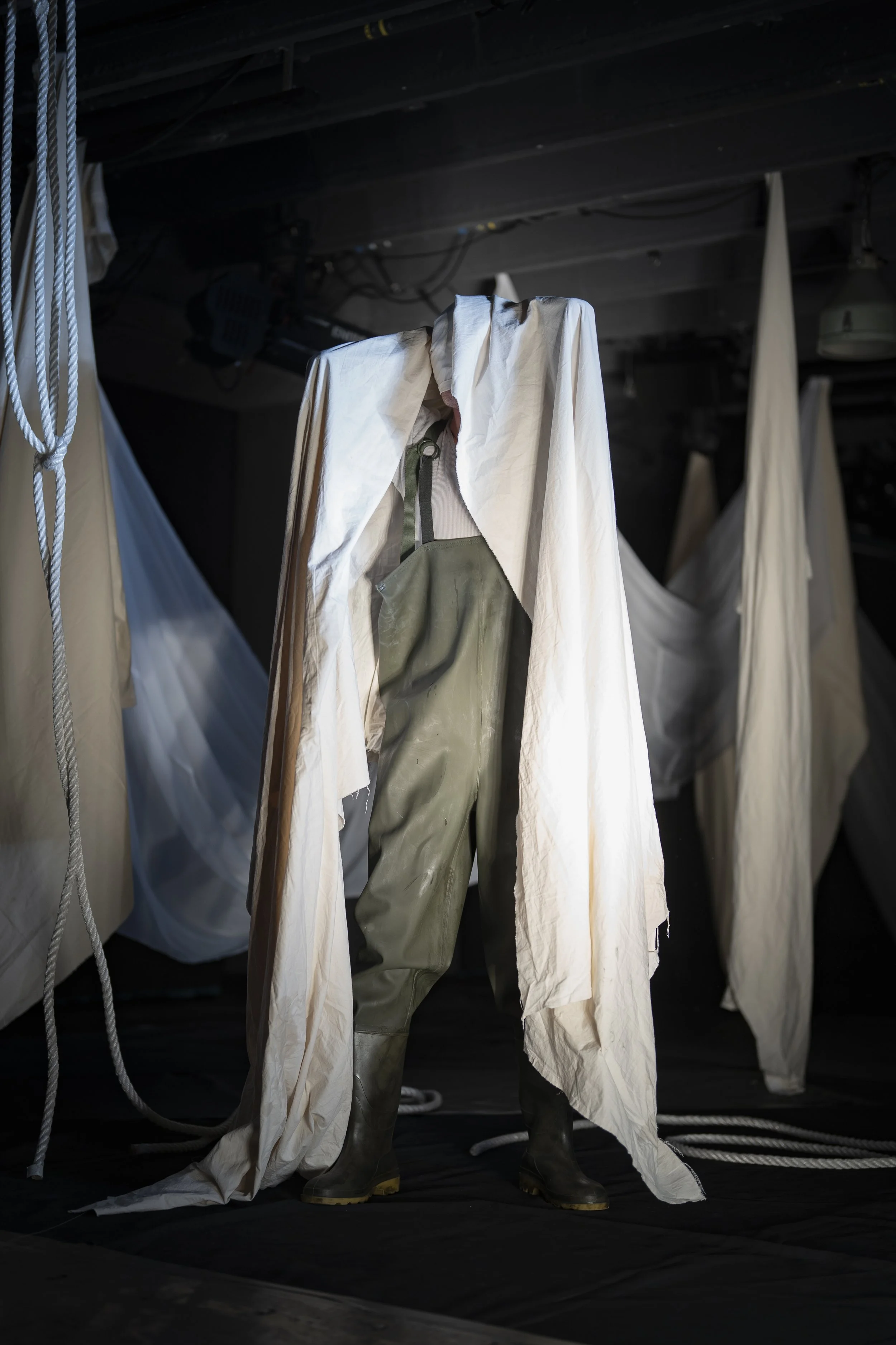 A person wearing green rubber boots, beige overalls, and a white protective suit stands among hanging white fabric and ropes in a dark environment. It is an image of Hugo Tiedje.
