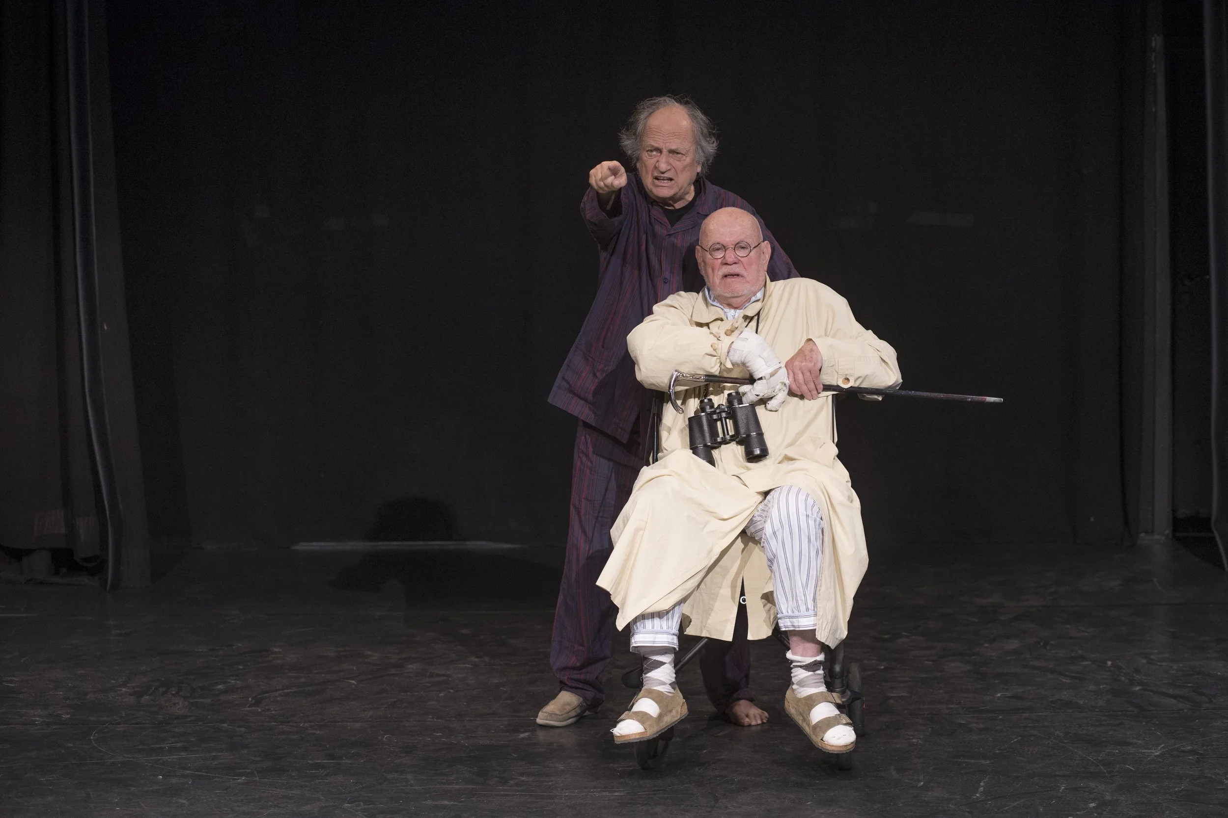 Two men performing on stage, one sitting in a wheelchair with binoculars, and the other standing behind him, pointing and speaking.