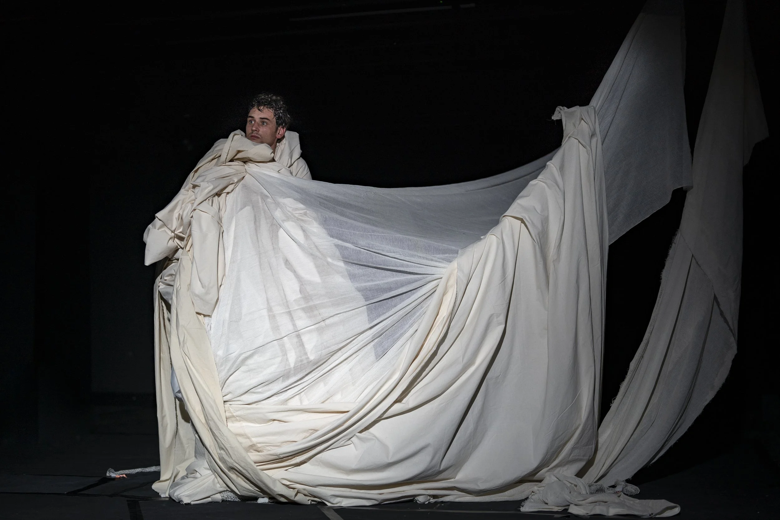 A man with curly hair bulked up in a large, white, draped fabric structure on a dark stage, looking upward. It is an image of Hugo Tiedje.