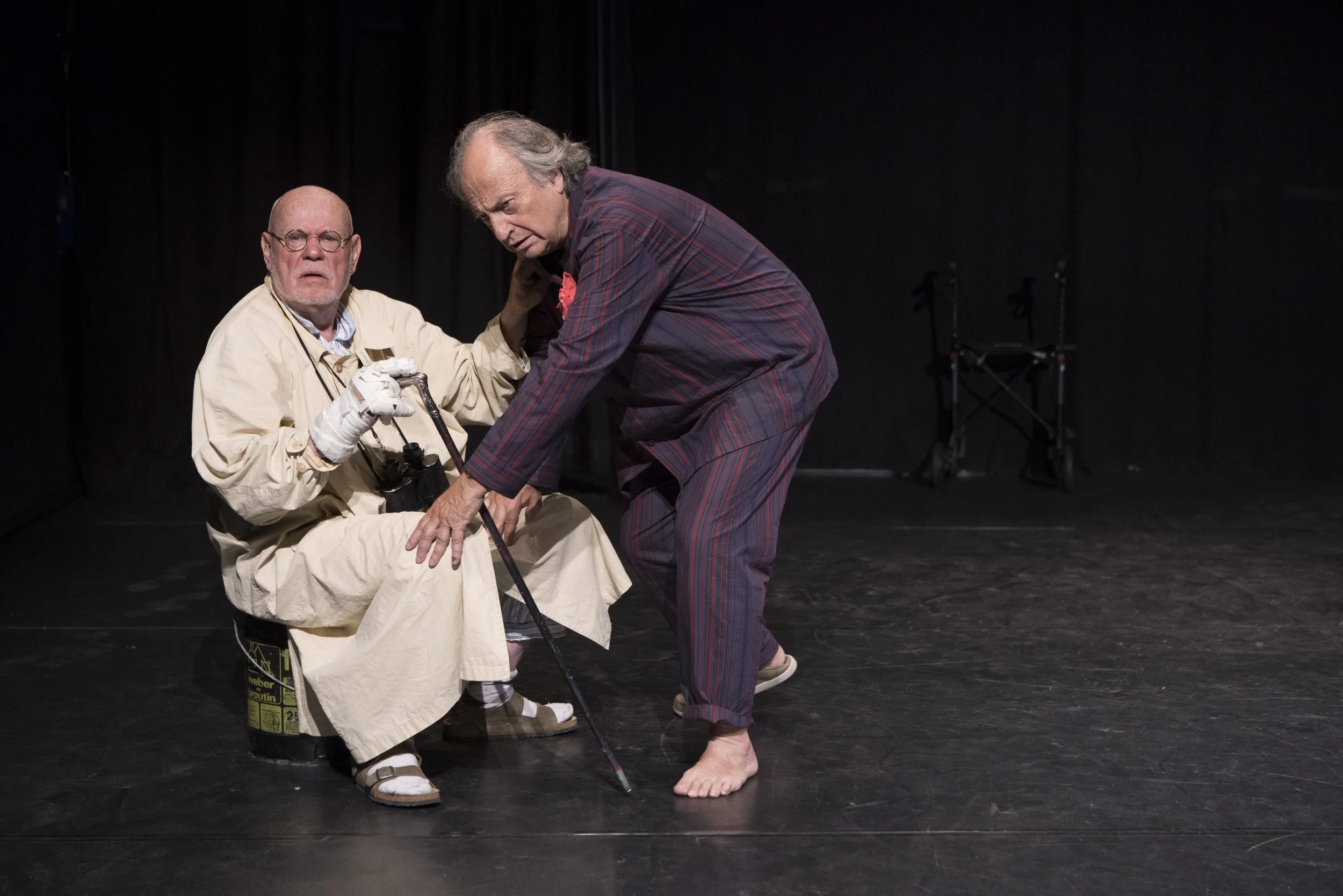 Two older men on a stage, one sitting with a cane and binoculars, the other leaning forward, both appearing to be engaged in a theatrical performance with a black background and minimal props.