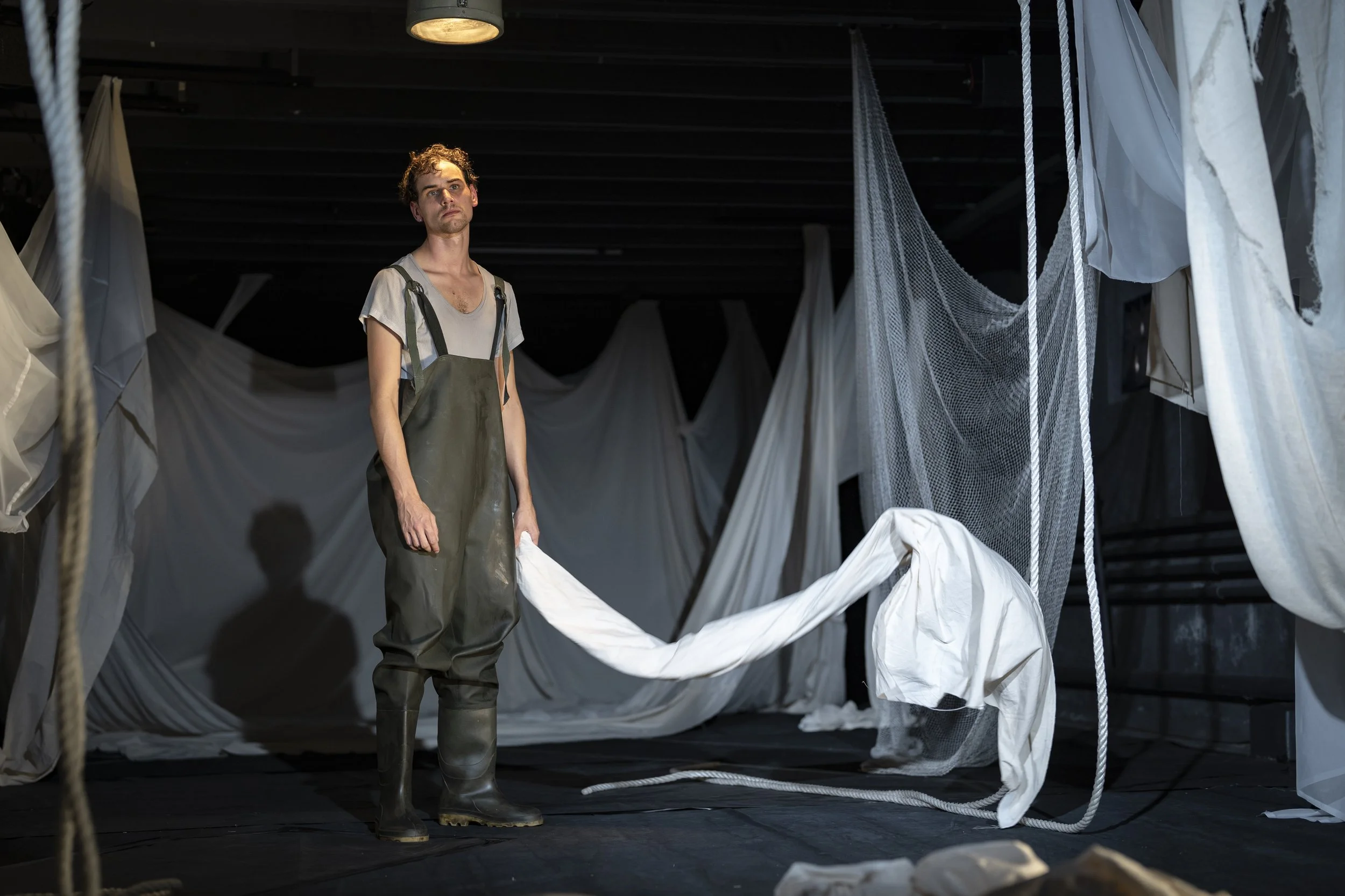 A young man stands in a dimly lit room surrounded by white fabric sheets and hanging nets, wearing waterproof boots, rabbit-like overalls, and a casual t-shirt, gazing thoughtfully into the distance. It is an image of Hugo Tiedje.