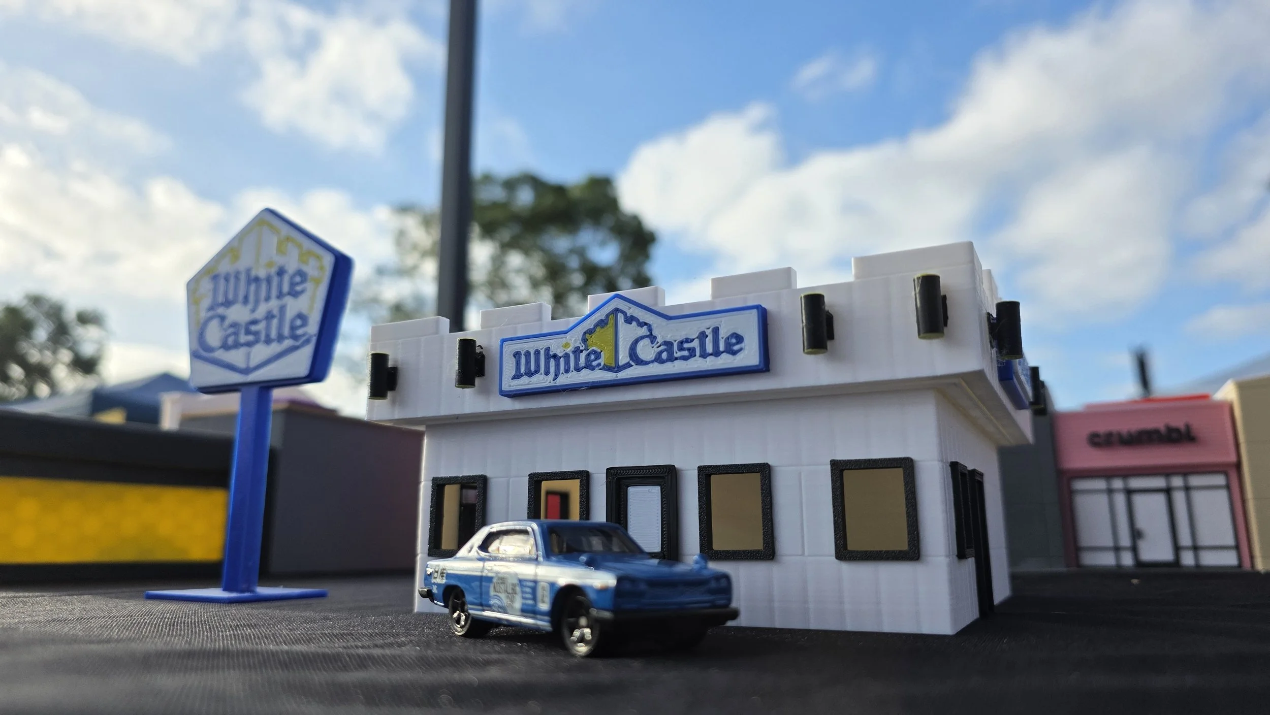 A small model of a White Castle restaurant with a White Castle sign, a toy car, and a blue flag, set outdoors against a background of trees and sky with clouds.