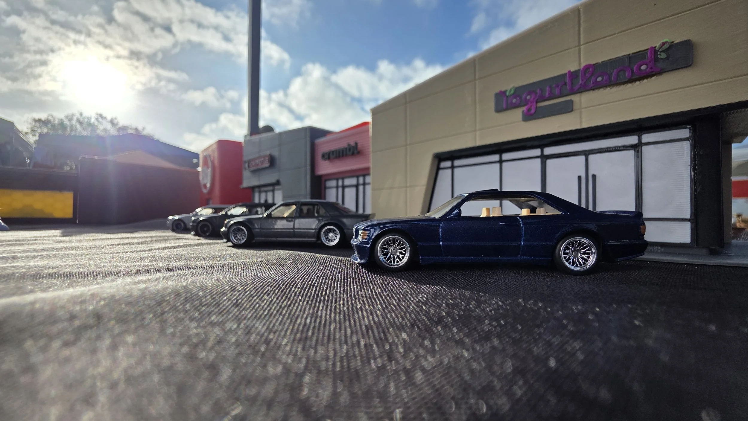 Small toy cars parked in front of a shopping plaza with store signs for 'iegurtland', 'crook', and 'grombi'. The photo is taken outdoors with sunlight and partly cloudy sky.