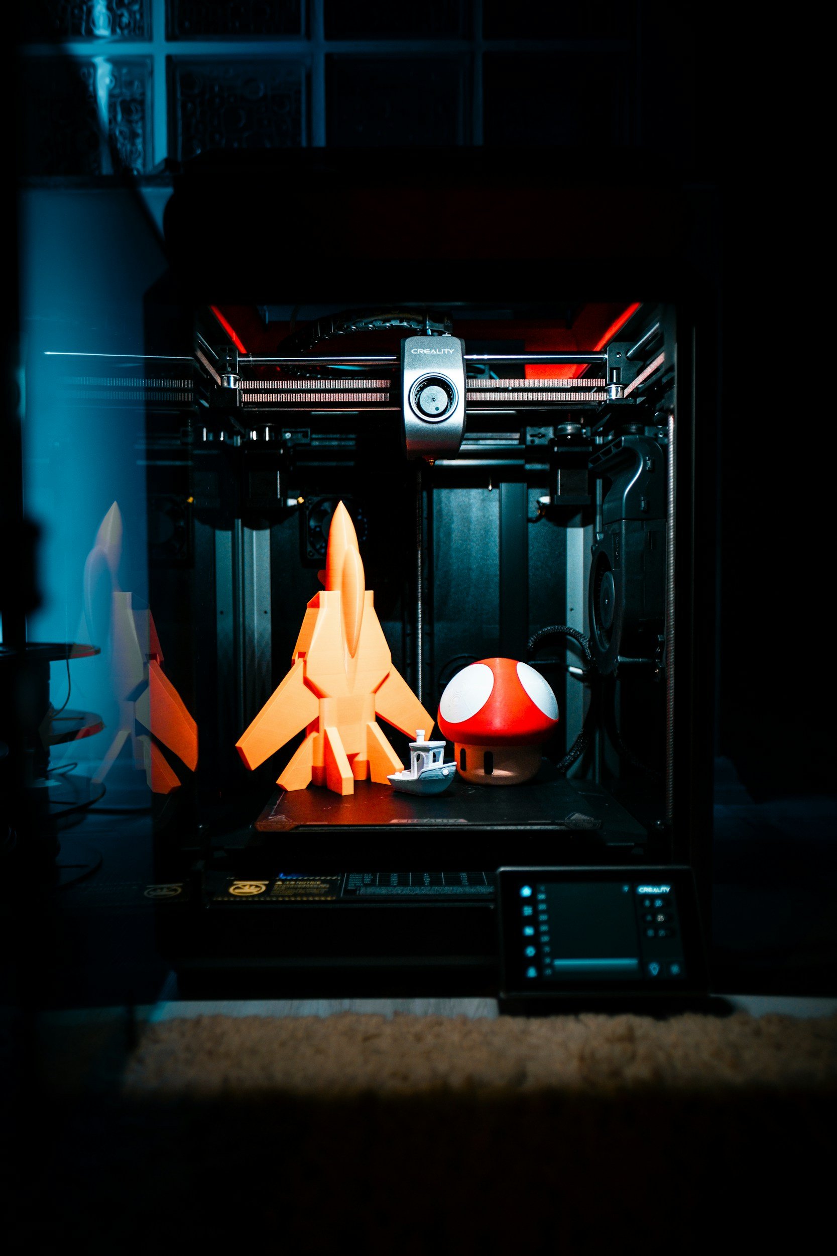 Image shows a 3D printer in action with a toy rocket, ball mushroom, and a small boat-like object on its printing bed, in a dimly lit room with a glass block wall in the background.
