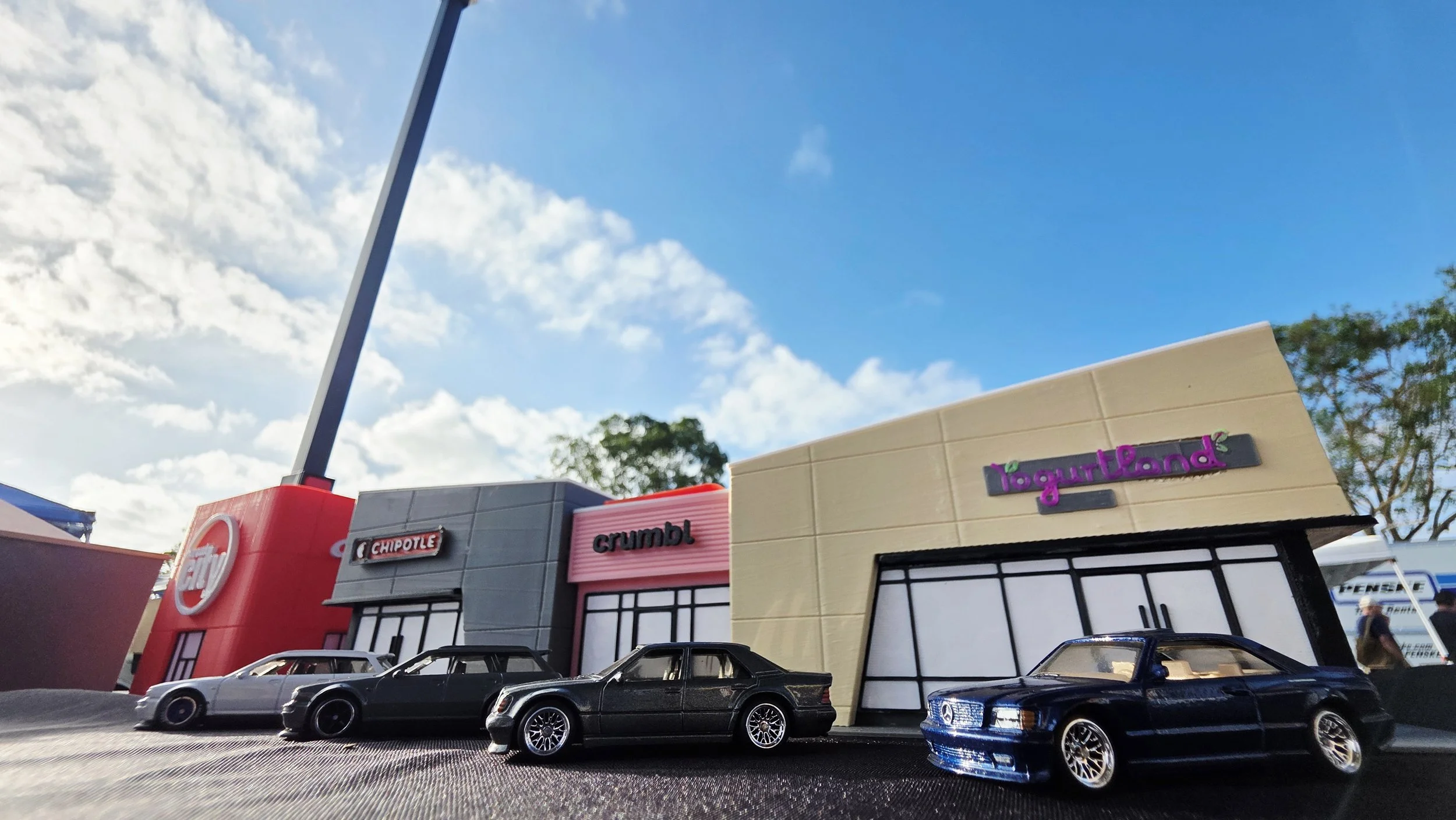 A shopping mall parking lot with toy cars in the foreground and a strip mall in the background, featuring a Retail Store, Chipotle, Crumbl, and LogitLand storefronts, under a partly cloudy blue sky.