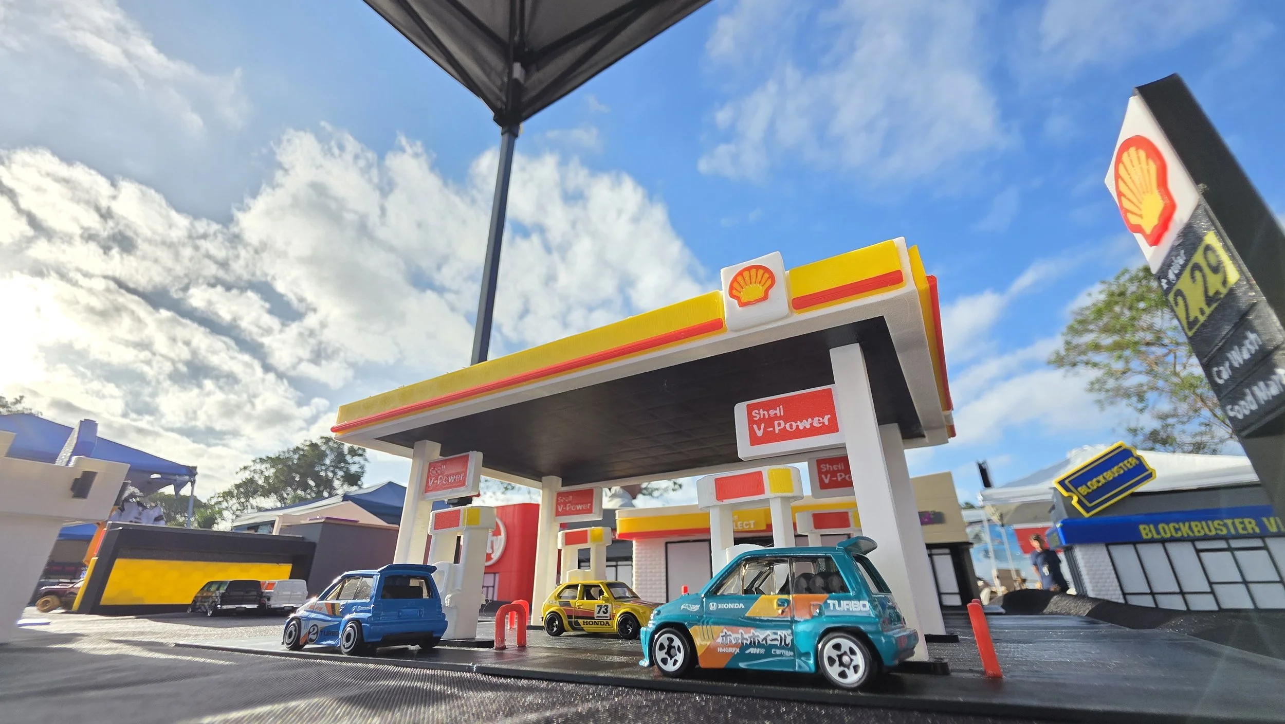 Toy gas station with toy cars parked in front, featuring Shell branding and a clear sky with clouds in the background.