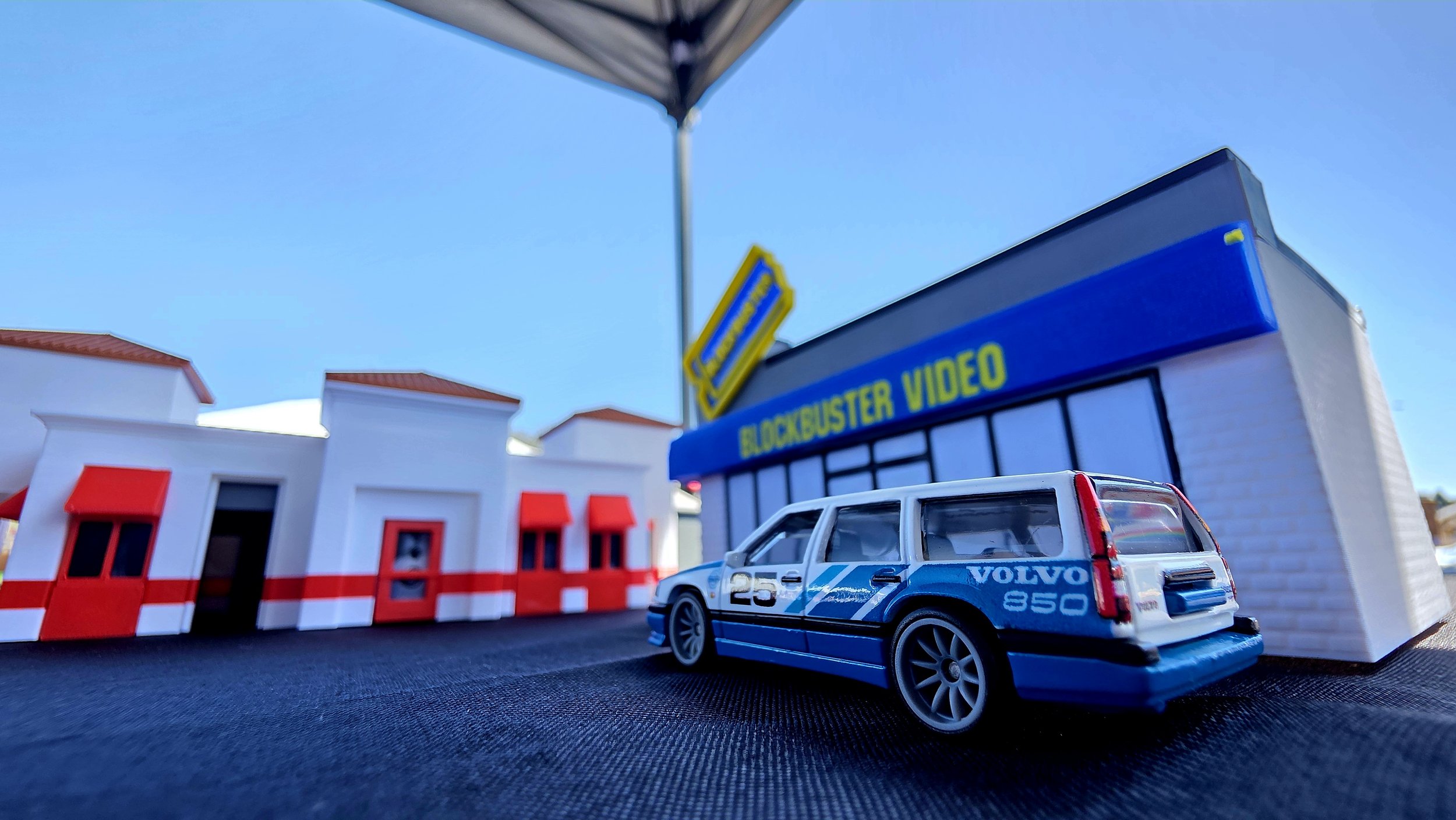 Miniature model of a blue Volvo 850 station wagon with racing decals parked in front of a blocky building resembling Blockbuster Video store under a clear blue sky.