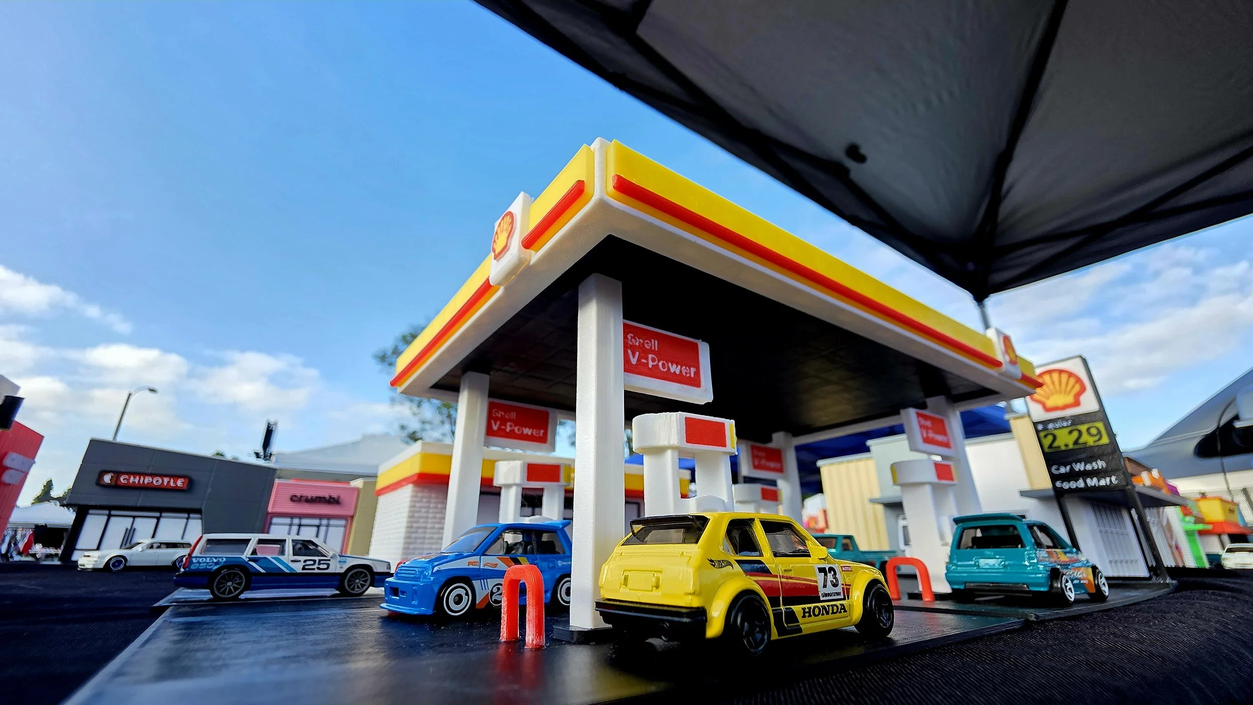 Miniature toy cars and a toy gas station set up as a display, with a Shell gas station logo and buildings in the background.