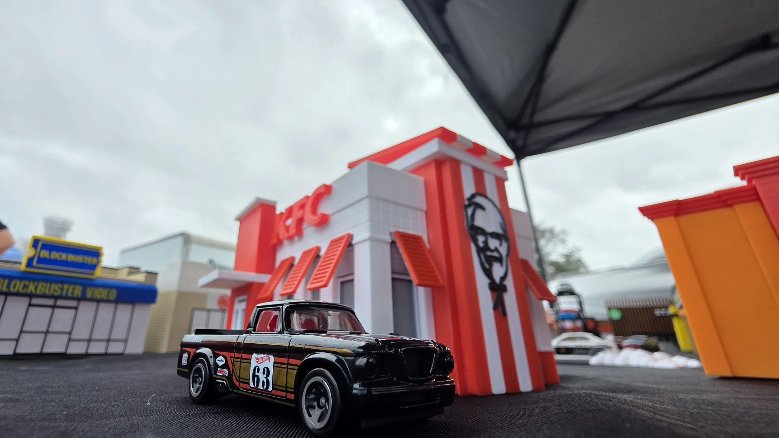 A small toy model of a black race car with racing decals, number 63, on a miniature city street scene with fast food restaurant buildings and a cloudy sky.
