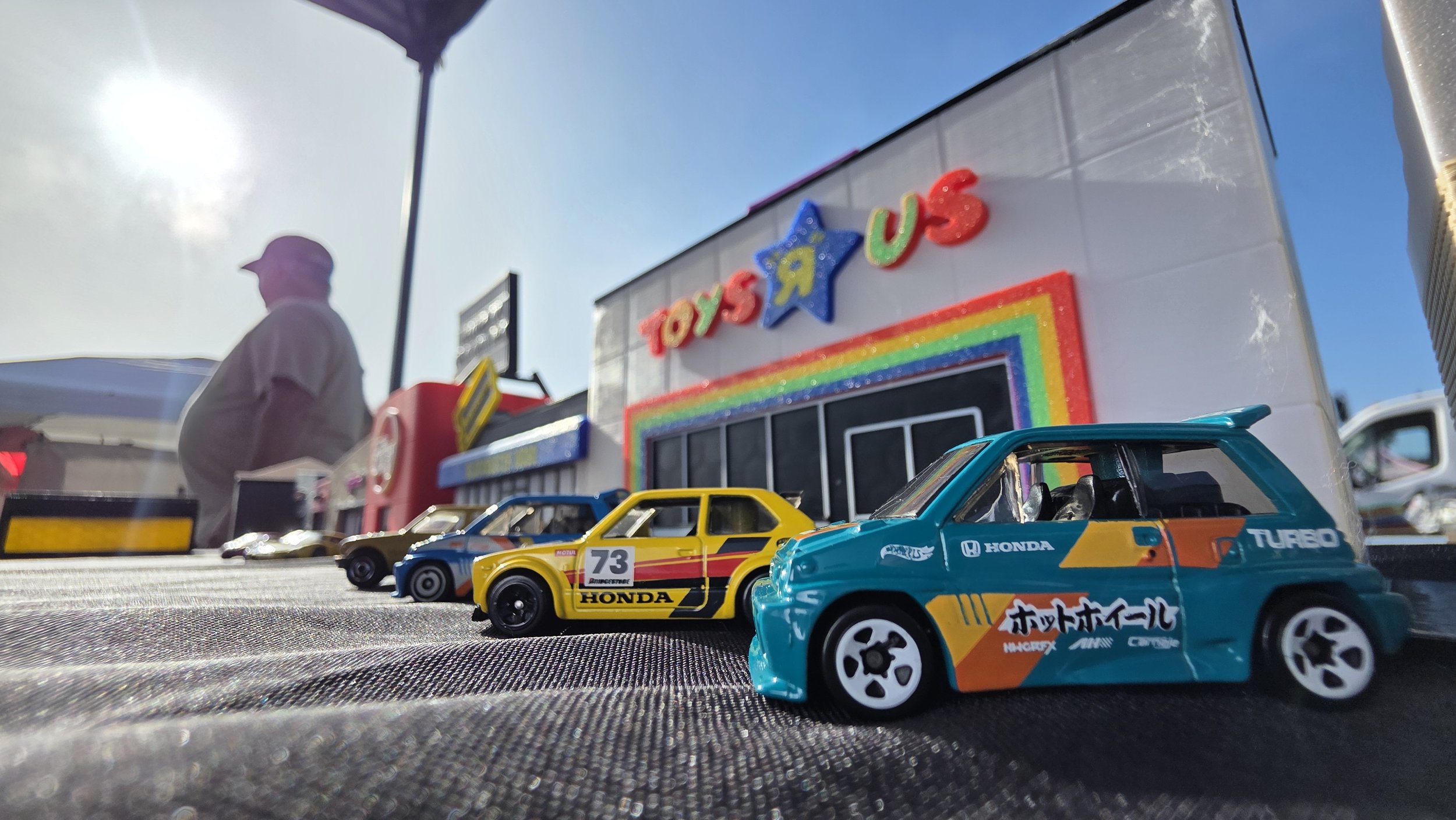 Close-up of miniature race cars on a textured surface, with a building decorated for Toys R Us in the background. The building features bright rainbow colors and a large blue star with the words 'TOYS R US'.
