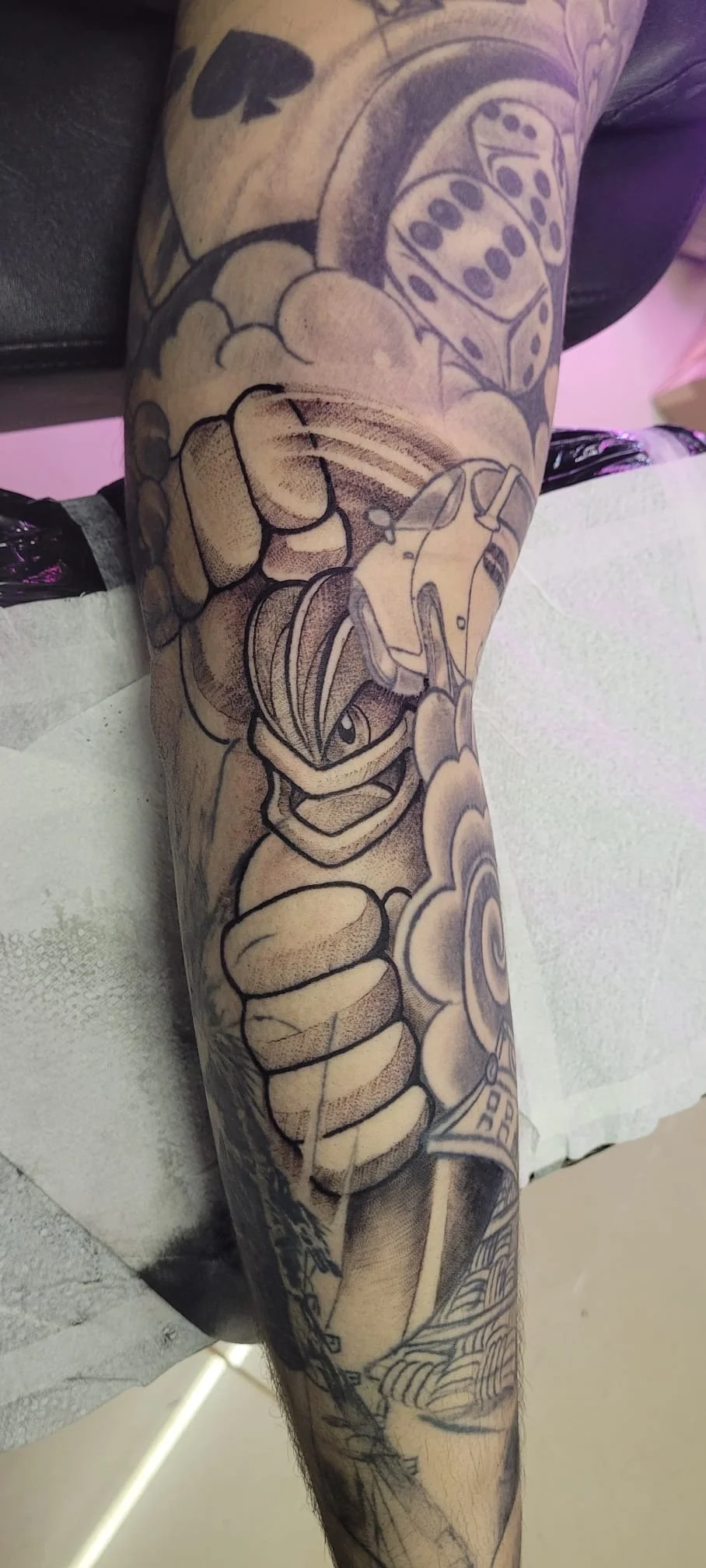 Black-and-gray cartoon character tattoo sleeve featuring dice, flowers, and a stylized masked figure on a person’s arm