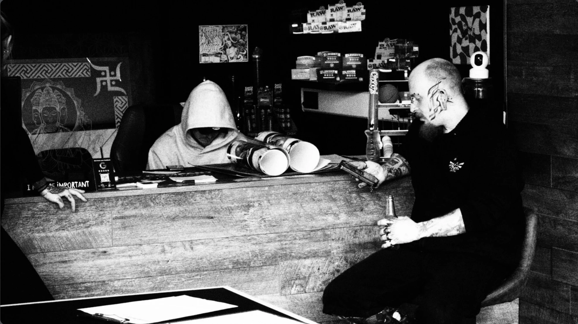 Black-and-white photo of a tattooed man holding a beer beside a hooded person seated behind a cluttered bar counter.