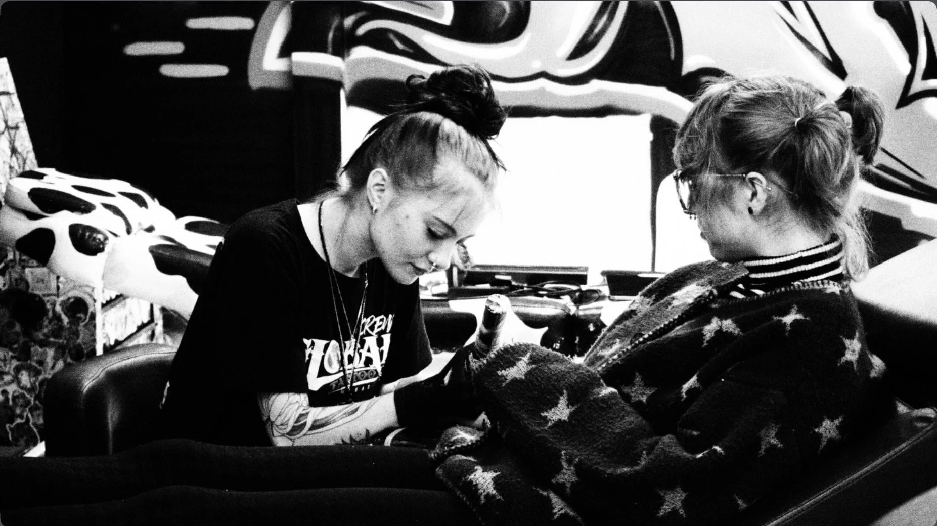 Black-and-white photo of two women sitting together indoors, one wearing glasses and a star-patterned sweater.