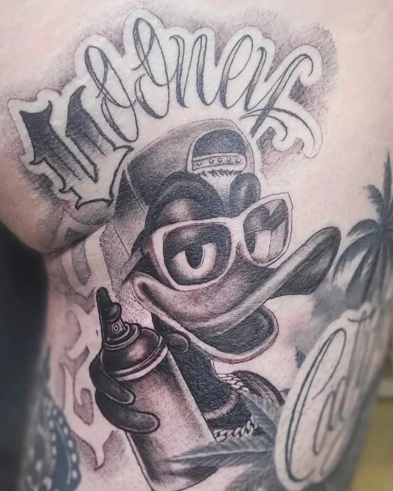 Black-and-gray tattoo of a graffiti-style cartoon duck wearing sunglasses and a backward cap, holding a spray paint can, with lettering above.
