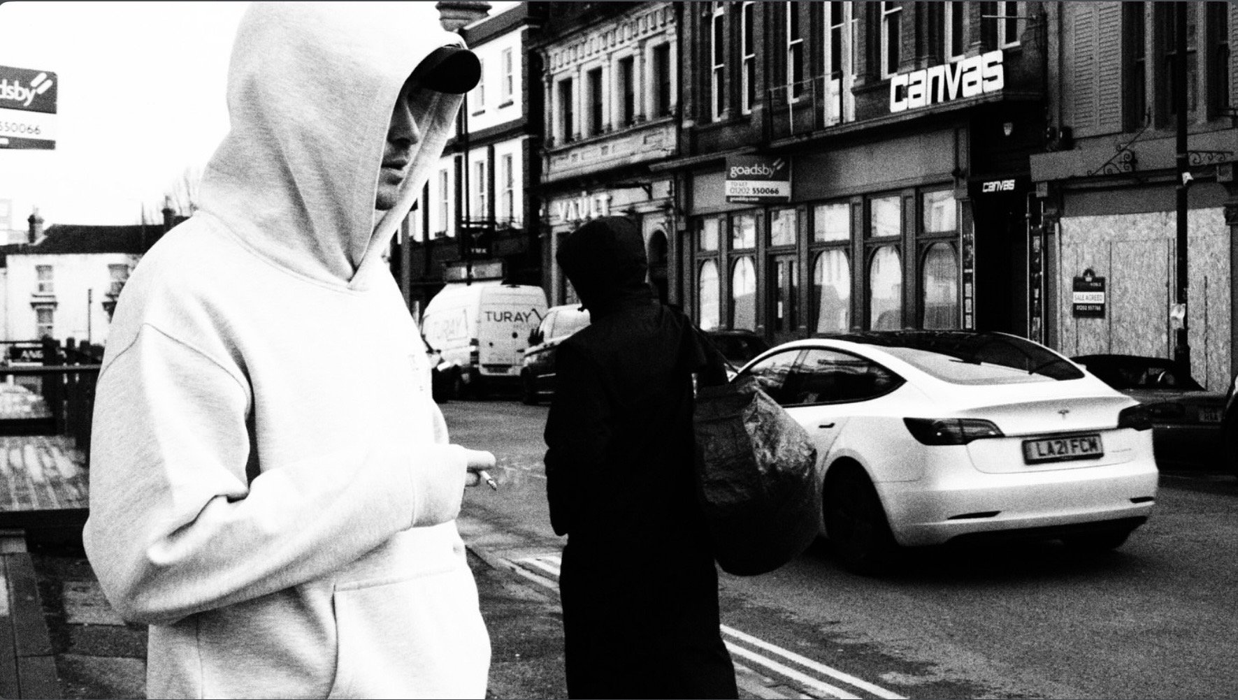 Black-and-white street scene with a hooded person, pedestrian, storefronts, and a parked white Tesla.