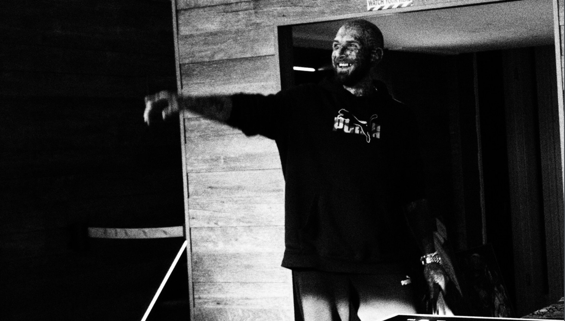 Black-and-white photo of a smiling bearded man in a Puma sweatshirt standing in a doorway with one arm extended.