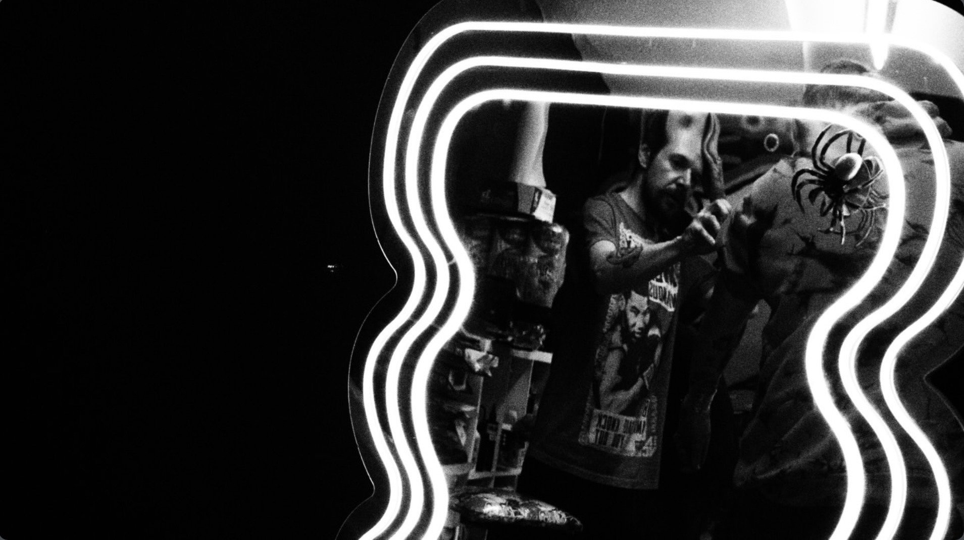 Black-and-white reflection of a tattooed person taking a mirror selfie beside glowing wavy neon lights
