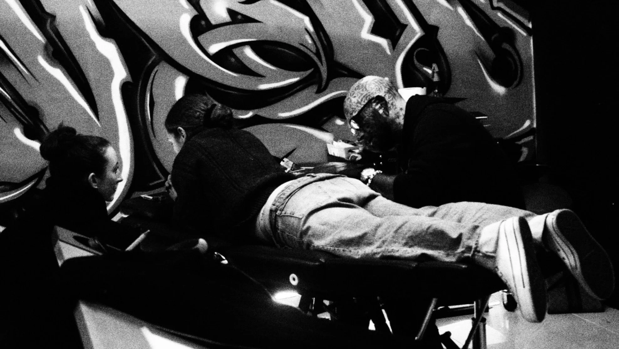 Black-and-white photo of three people gathered around a person lying on a stretcher at an indoor event.