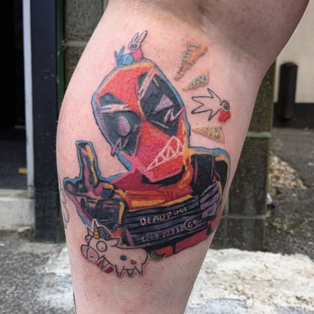 Colorful tattoo of Deadpool riding a motorcycle, with small cartoon figures and symbols around the design on a person’s thigh