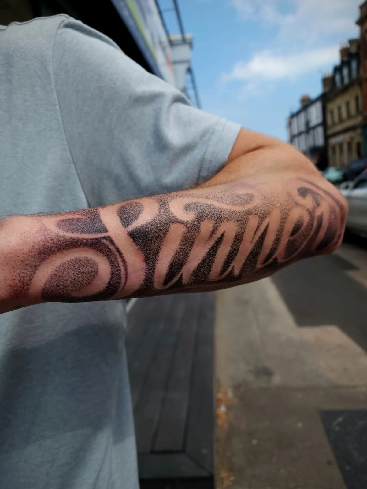 Close-up of a person's tattooed forearm in a gray shirt on a city street