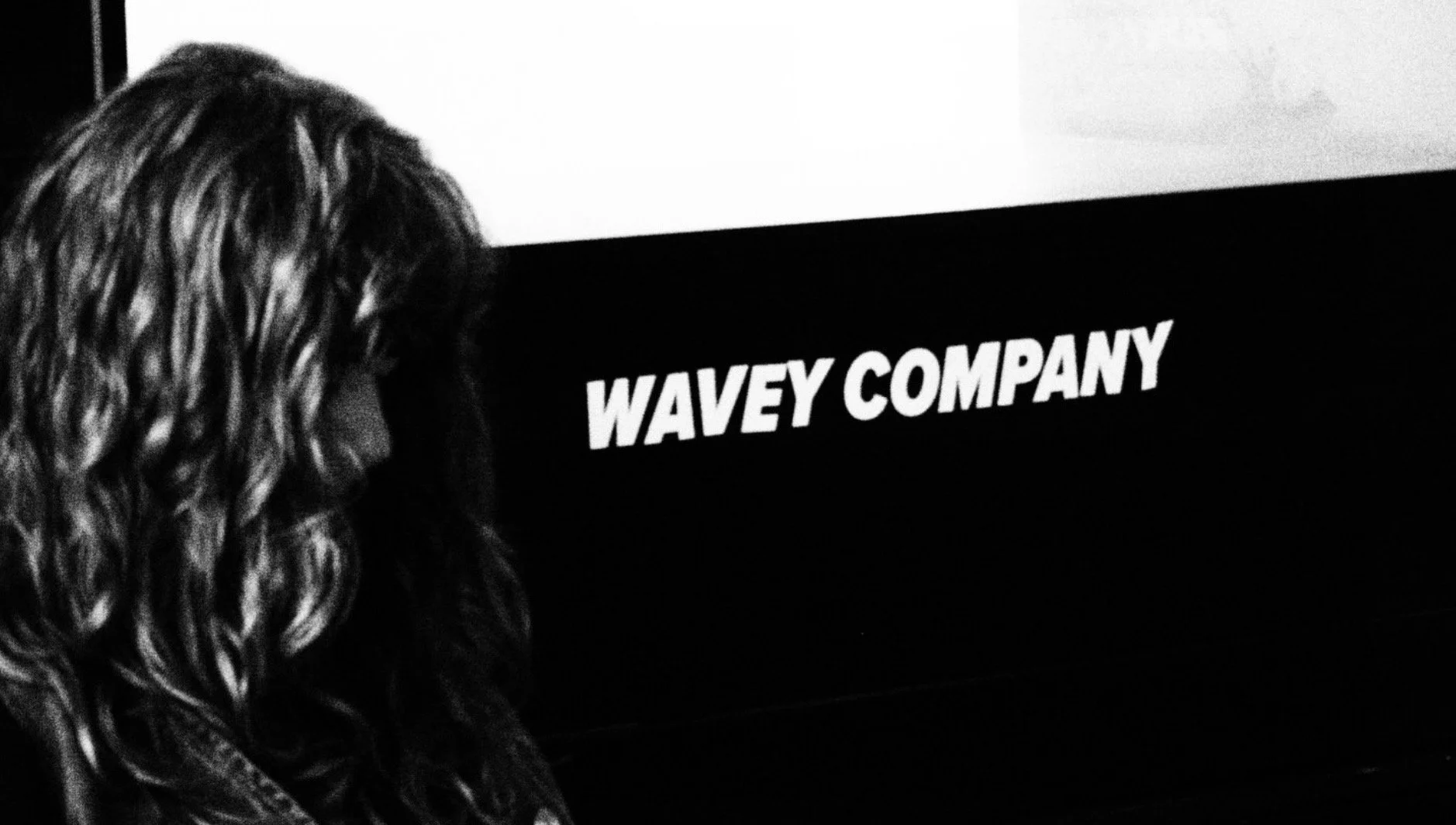 Black-and-white image of a person with long wavy hair beside a sign reading “WAVEY COMPANY.”
