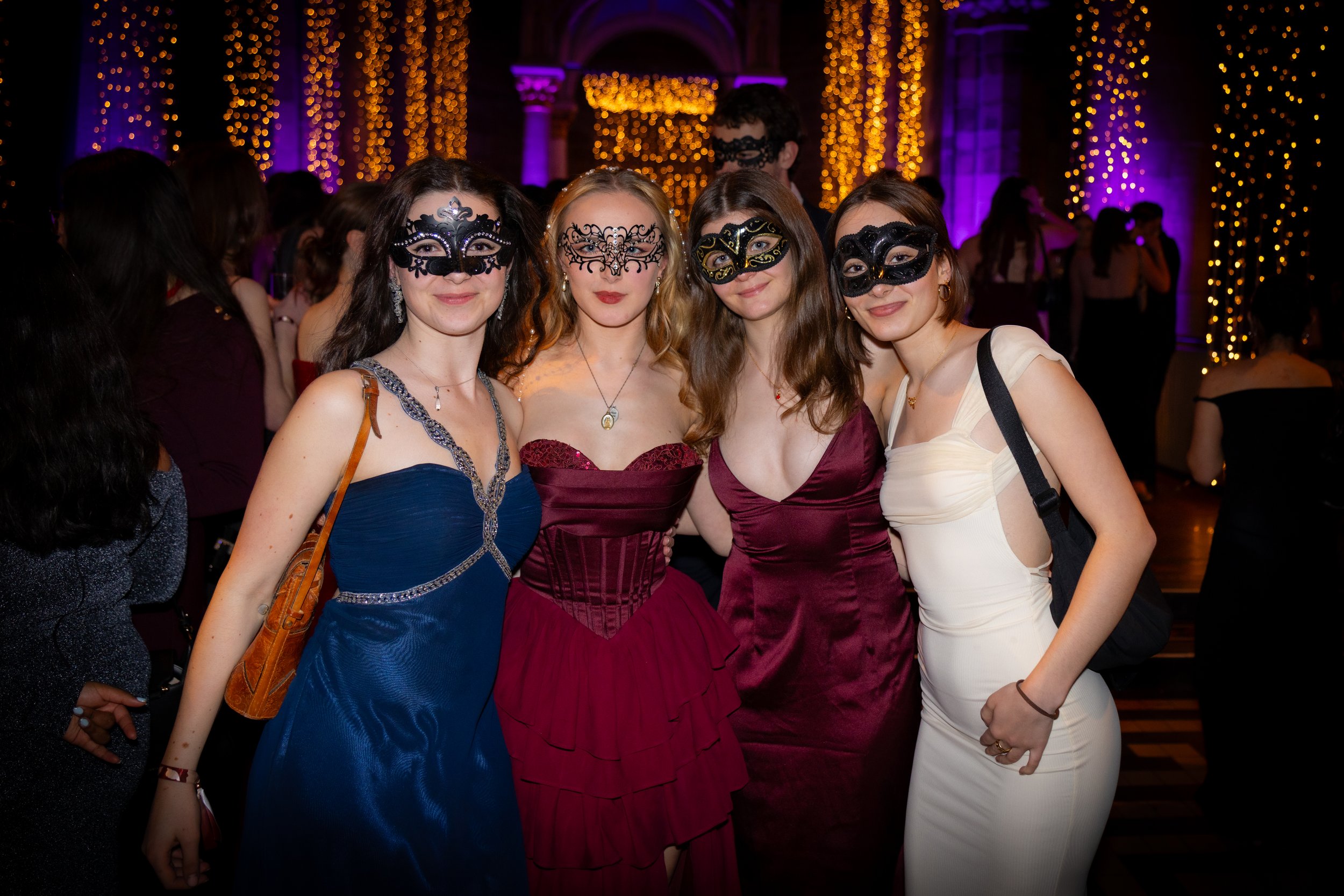 Four young women wearing elegant dresses and decorative masquerade masks standing together at a festive event with purple and orange string lights in the background.