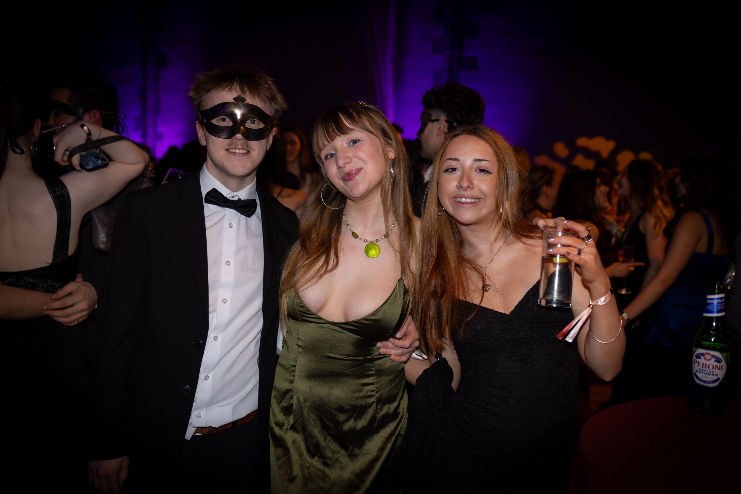 Three young adults at a party smiling for the camera. The man on the left is wearing a tuxedo, bow tie, and a black mask. The woman in the middle has long red hair, earrings, and a green dress. The woman on the right has long red hair, a nose ring, a