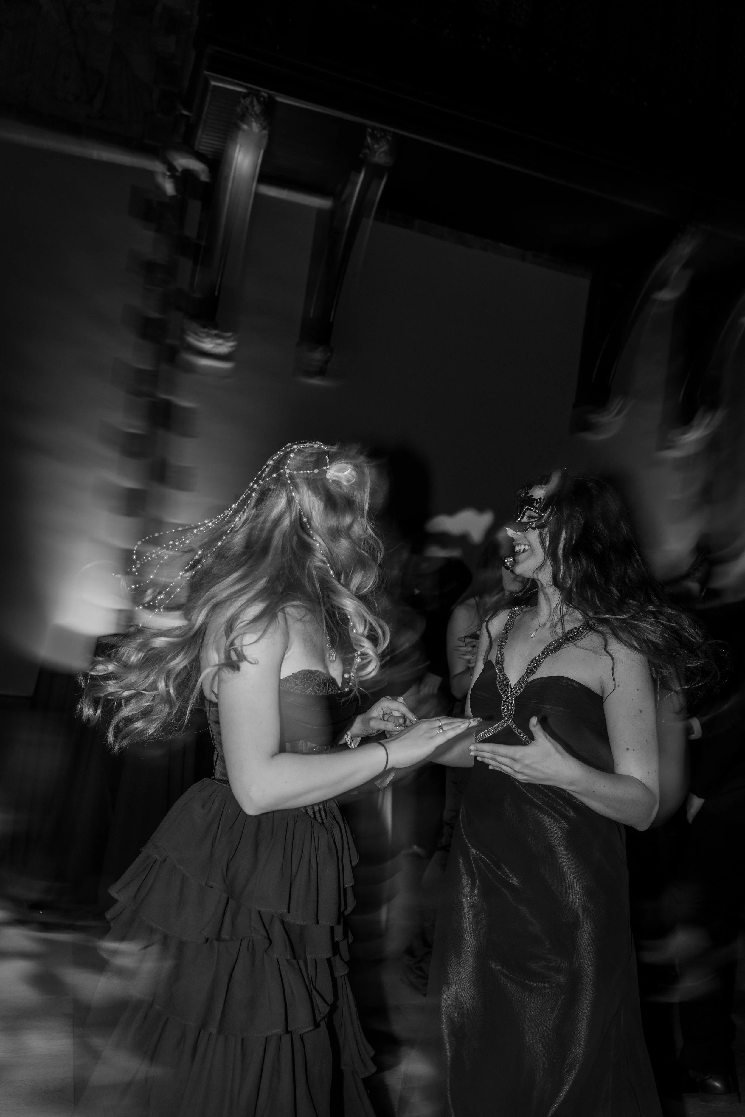 Two women in dresses dancing and smiling at a party or celebration, with a dark background and musical equipment visible above.